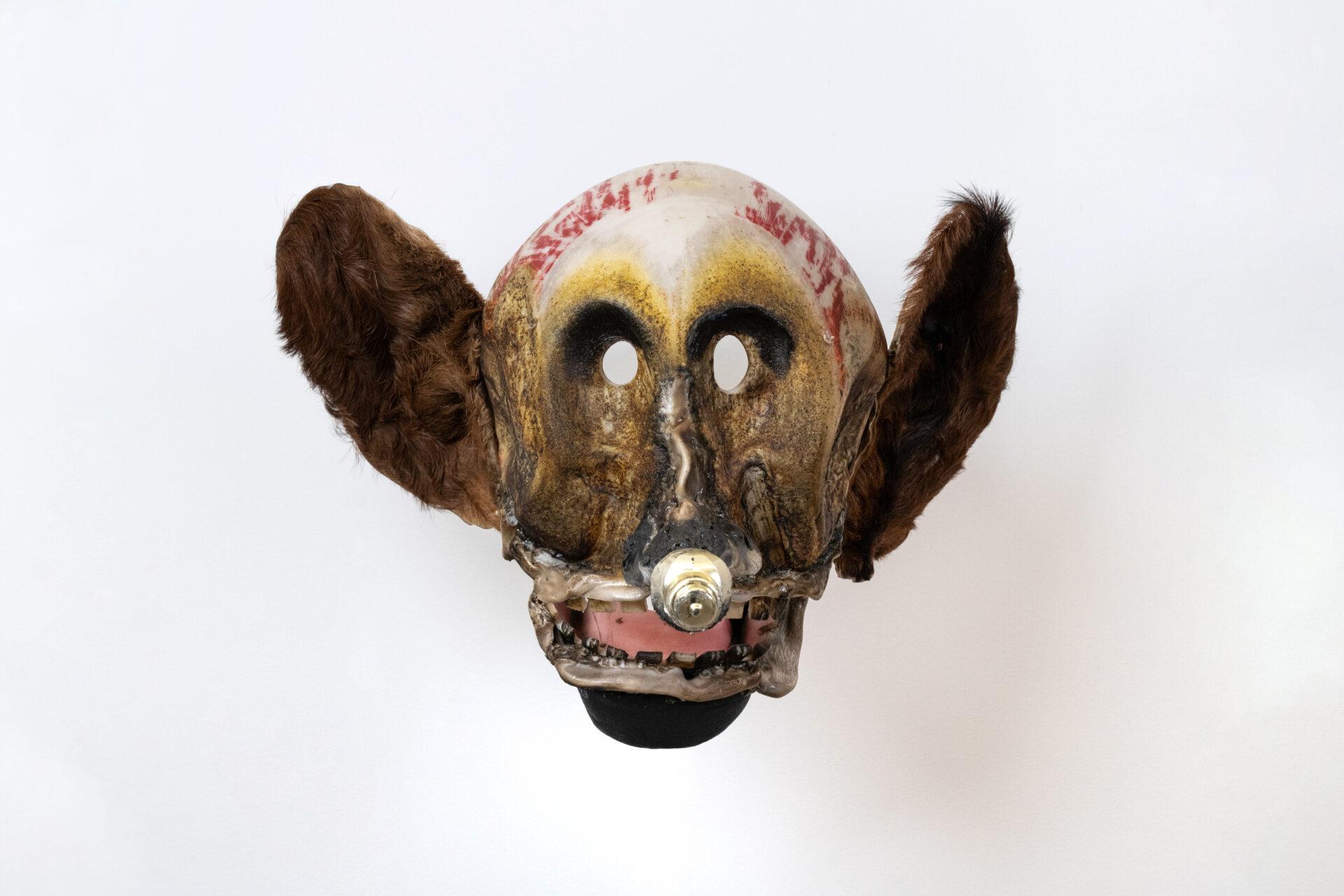 Descrição da imagem: Máscara tridimensional pintada. Vista de frente, a obra é arredondada, com o topo da cabeça em bege manchado de vermelho. As orelhas são grandes, arredondadas e cobertas por pêlos marrons. Os olhos são ovais e vazados, com a área superior pintada de preto.
O nariz é alongado, em marrom e preto, com a ponta arredondada projetada para frente. A boca é larga e está aberta, com lábios beges e grossos. Os dentes são pequenos, quadrados, brancos, com manchas pretas, e a língua é rosa. O queixo é preto. Fim da descrição da imagem.
