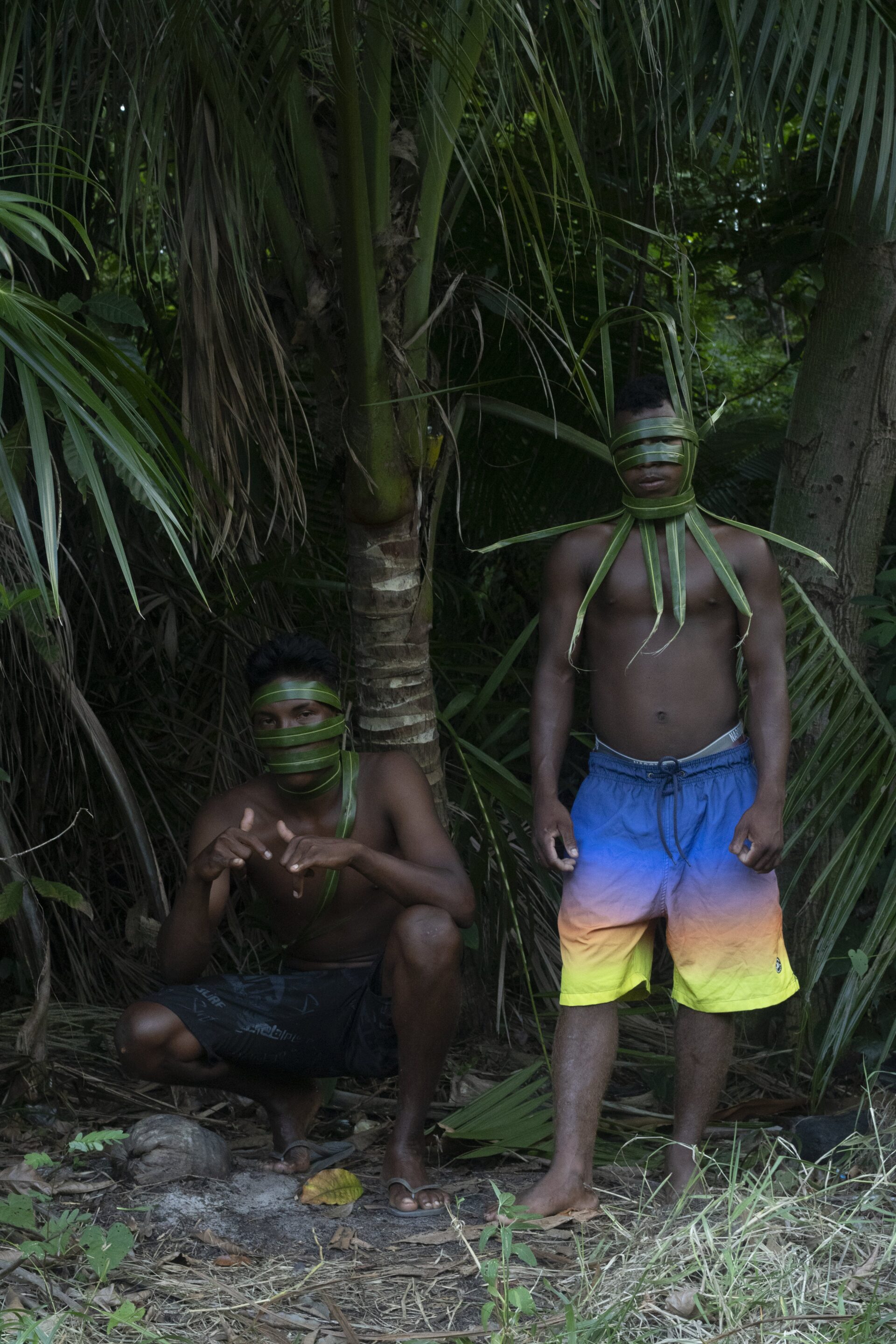 Descrição da imagem: Fotografia diurna, em formato vertical, de dois homens em um ambiente natural com vegetação densa composta por palmeiras e folhagens verdes.
Os dois homens são negros e estão diante das palmeiras.
O homem da esquerda está agachado, com o tronco levemente inclinado para frente e os dedos polegar e mindinho de ambas as mãos erguidos. Tem cabelos pretos em topete e sobrancelhas finas. Folhas de palmeira envolvem o rosto em faixas que deixam apenas os olhos escuros e o queixo à mostra. O peito está nu e a bermuda é azul-escura com estampa.
O homem da esquerda está agachado
O homem da direita está em pé, com os braços ao lado do corpo. O cabelo é preto, o bigode ralo e os lábios grossos. Folhas de palmeira envolvem o rosto em faixas que deixam apenas os olhos escuros e a parte da boca à mostra. Uma faixa de folha de palmeira está em torno do pescoço, sustentando outras folhas dispostas para cima, para baixo e para os lados. O peito está nu e a bermuda é em degradê de azul, rosa, laranja e amarelo. Fim da descrição da imagem. 
