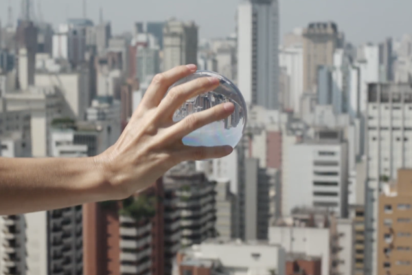 Imagem. Descrição da imagem: Em primeiro plano, uma mão de pele clara segura uma esfera lisa e transparente, que parece ser de vidro ou cristal. A mão está posicionada da esquerda para o centro da imagem e a esfera reflete e refrata a paisagem ao fundo de ponta-cabeça. Ao fundo, sob luz solar clara, estende-se a vista de uma grande cidade com muitos arranha-céus, em uma paleta de cores neutras e tons de cinza. O céu está ligeiramente esbranquiçado, com a cena iluminada por luz natural de um dia claro. A iluminação destaca a transparência e o efeito de lente da esfera. Fim da descrição.