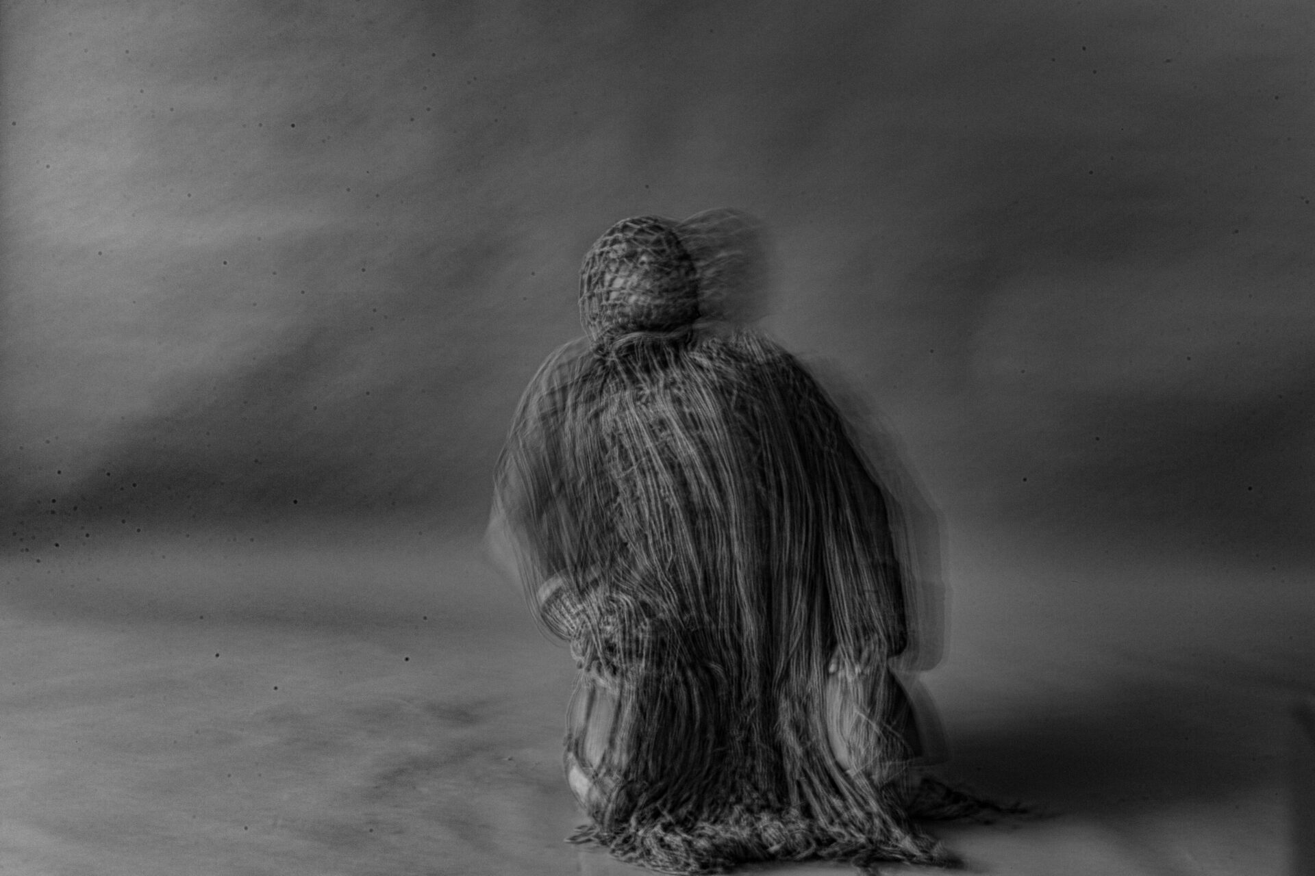 Descrição da imagem: Fotografia horizontal, em tons de cinza, desfocada. Ao centro, uma pessoa ajoelhada é vista de frente, com as mãos apoiadas nas coxas e o tronco levemente inclinado para trás. Ela está coberta da cabeça aos pés por uma vestimenta longa em tons de cinza-claro e cinza-escuro, formada por múltiplas tiras finas: no rosto, entrelaçadas em pequenos losangos; no corpo, dispostas verticalmente. Fim da descrição da imagem.

