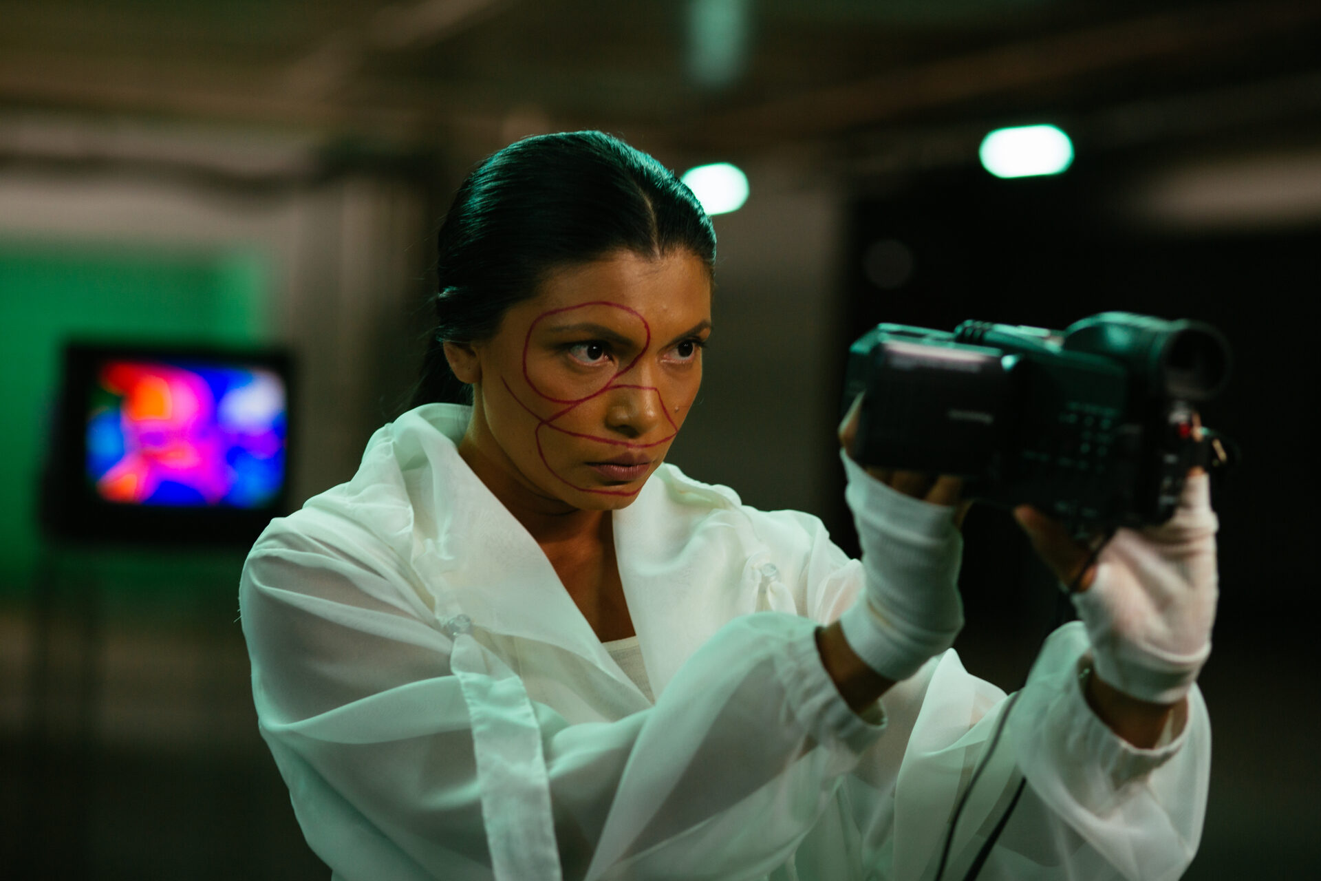 Descrição da imagem: Fotografia horizontal de uma mulher indígena em ambiente interno, vista do busto para cima. Tem a pele escura, cabelos pretos e lisos presos para trás, sobrancelhas finas, olhos escuros, nariz arredondado na ponta e lábios médios. Uma linha fina e vermelha está pintada em padrões circulares e curvos, contornando o olho direito, o nariz e a bochecha direita. Ela usa jaqueta branca translúcida e luvas brancas e segura com as mãos uma câmera de vídeo preta voltada para o próprio rosto. O fundo está desfocado. Fim da descrição da imagem.
