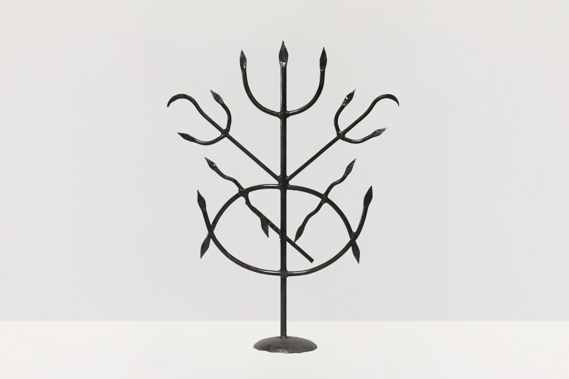 Descrição da imagem: Ferramenta Escrava de Iansã. A peça é sustentada por uma base circular, apoiada sobre chão claro.
Estrutura metálica vertical, escura, composta por hastes de ferro. Uma haste reta e central termina, no topo, em um tridente com três pontas afiadas.
No meio da haste central, duas hastes posicionadas em diagonal se estendem para as laterais; cada uma tem tridentes curvos nas extremidades, voltados para fora, e a ponta central curvada para baixo.
Abaixo dessas hastes, há uma haste curvada para baixo, com as pontas afiadas. Nela, à esquerda, uma haste representa uma lança ondulada com uma folha na parte central; à direita, outra lança ondulada com as extremidades pontudas.
Na parte inferior, há uma haste curvada para cima, cujas pontas se unem às pontas da outra haste curvada, formando uma figura oval. Fim da descrição da imagem.
