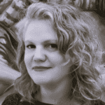 Imagem. Início da descrição de imagem: Retrato em close-up em tons de sépia ou preto e branco de uma mulher de pele clara e cabelos loiros ou grisalhos, ondulados e volumosos, que emolduram seu rosto. Ela tem uma expressão suave, com os lábios levemente marcados, e veste uma peça de roupa escura. O fundo está desfocado, com texturas indistintas. Fim da descrição de imagem.