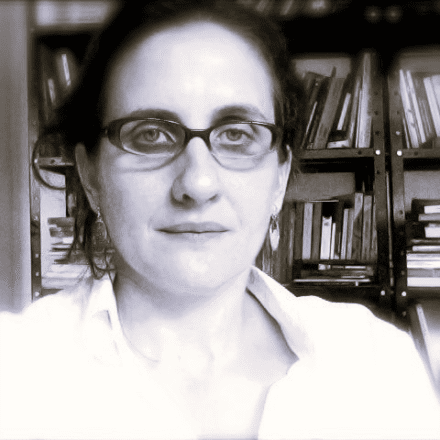 Imagem. Início da descrição de imagem: Retrato em tons de sépia ou preto e branco de uma mulher de pele clara, com cabelos escuros presos e usando óculos de grau de aro preto retangular. Ela veste uma camisa social branca e olha diretamente para a câmera com uma expressão neutra. Ela está posicionada em frente a estantes de livros escuras e repletas de volumes, que servem como fundo para o retrato. Fim da descrição de imagem.