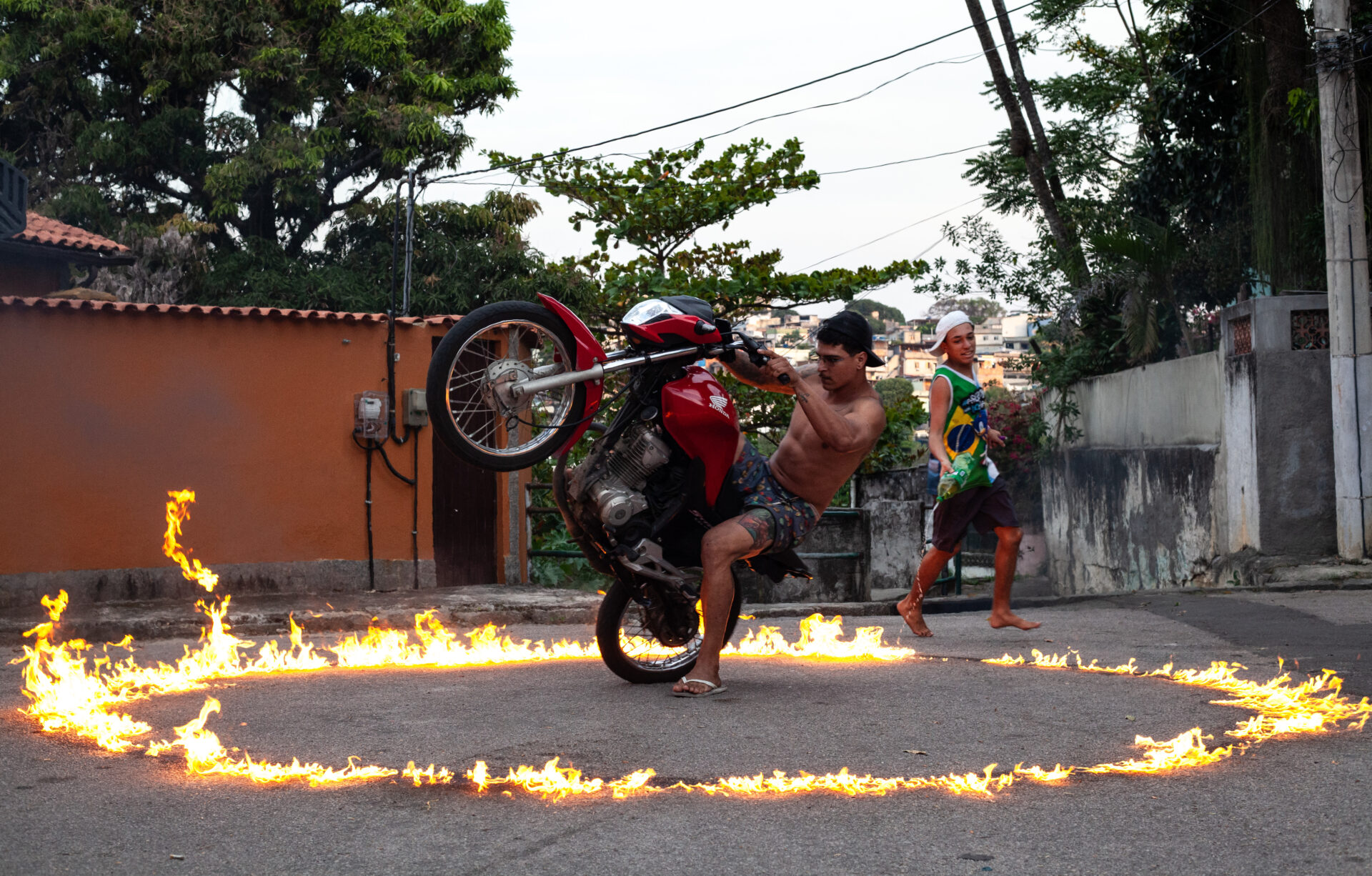 Descrição da imagem: Fotografia horizontal. Sob céu cinza, em uma rua asfaltada, há um homem em uma moto vermelha com a roda dianteira empinada para a esquerda. Ele segura o guidão com as mãos. Ele é pardo, usa boné preto de aba curva virado para trás, óculos de grau de aros retangulares, está sem camisa, de bermuda escura estampada e chinelos brancos.
No chão, ao redor da moto, um grande círculo de chamas baixas em tons de amarelo-laranjado.
À direita do círculo, há um jovem pardo com boné branco de aba curva virado para trás, regata verde estampada com a bandeira do Brasil, bermuda escura e descalço. Ele segura uma grande garrafa PET, com a abertura voltada para baixo, da qual um líquido cai sobre as chamas.
Ao fundo, à esquerda, há um muro cor de laranja; à direita, um muro de concreto. Atrás deles, árvores com folhagens verdes. Fim da descrição da imagem.
