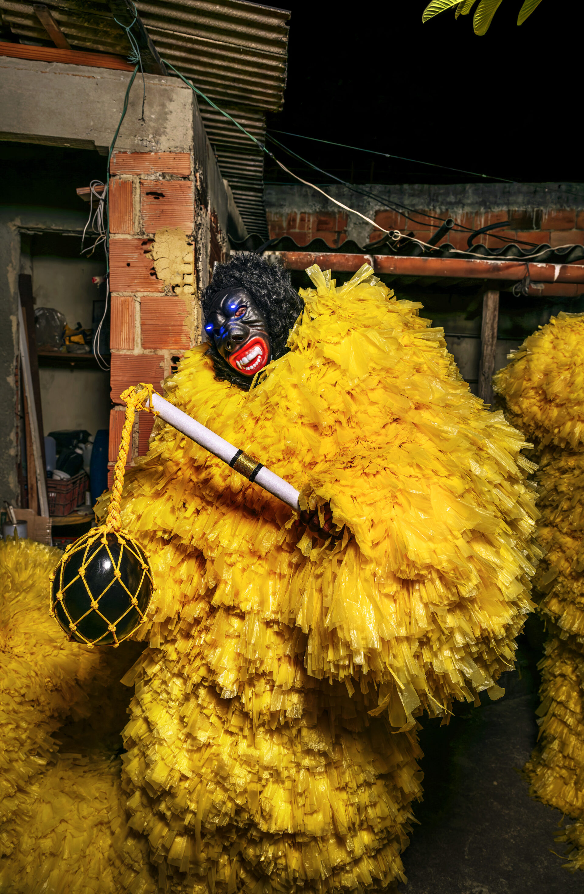 Descrição da imagem: Fotografia noturna, em formato vertical, de uma pessoa com indumentária de Gurilouko.
Está enquadrada dos joelhos para cima. A cabeça e o rosto estão ocultos por uma máscara semelhante à de um gorila.
A máscara tem cabelos curtos e pretos, pálpebras azuis, olhos vermelhos, nariz grande e boca vermelha, que está aberta, deixando os dentes brancos com caninos pontudos à mostra.
A roupa é amarela e bufante, semelhante a um macacão de mangas longas, feita de diversas tiras finas e compridas de sacolas plásticas.
A pessoa segura um bastão branco na diagonal para a esquerda; na ponta dele, uma corda amarela está amarrada e suspende uma bola de borracha preta coberta por uma rede amarela.
Ao fundo, há uma parede sem reboco, um pilar de concreto e, acima, um teto de telhas onduladas. Fim da descrição da imagem.
