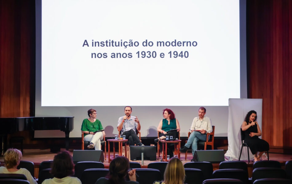 Descrição da imagem: Uma fotografia de um debate em um auditório com fundo de madeira e um grande telão. O telão exibe em fundo branco o texto "A instituição do moderno nos anos 1930 e 1940" em fonte escura. Quatro debatedores e uma intérprete de Libras estão sentados no palco. Um homem de camisa branca e uma mulher de camisa turquesa estão à esquerda. No centro, um homem com camisa clara está falando ao microfone. Uma mulher de blusa verde escura está sentada à sua direita. Na extrema esquerda, há uma mulher de roupa preta sentada em uma cadeira mais alta está gesticulando em Libras. A plateia no primeiro plano está vista de costas. Fim da descrição da imagem.