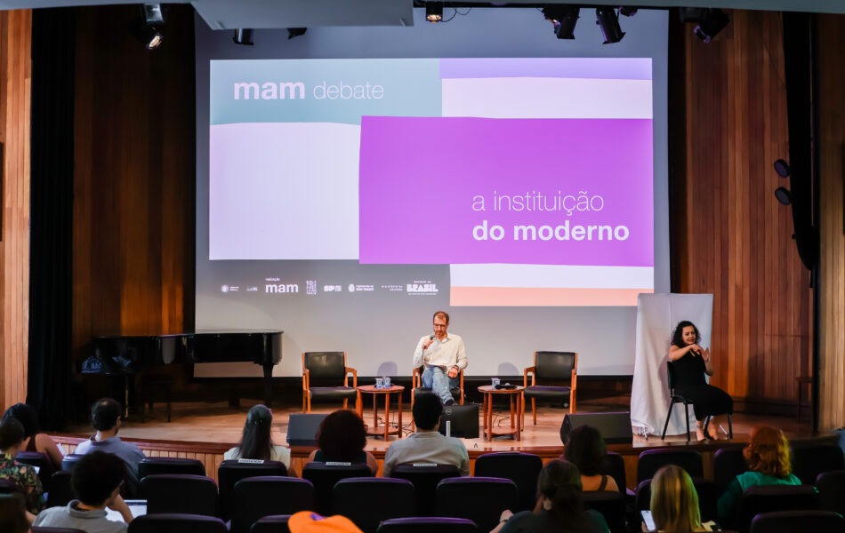Descrição da imagem: Uma fotografia de um debate em um auditório com fundo de madeira e um grande telão. O telão exibe o título "mam debate" e uma composição gráfica em branco, cinza e roxo com o texto "a instituição do moderno". Abaixo, estão logos de instituições. No palco de madeira, um homem no centro, com camisa branca e calça jeans, está sentado em uma cadeira escura, falando ao microfone. Ele está cercado por pequenas mesas redondas. À direita dele, uma intérprete de Libras, sentada em uma cadeira alta, gesticula. A plateia no primeiro plano está vista de costas em assentos escuros. Fim da descrição da imagem.