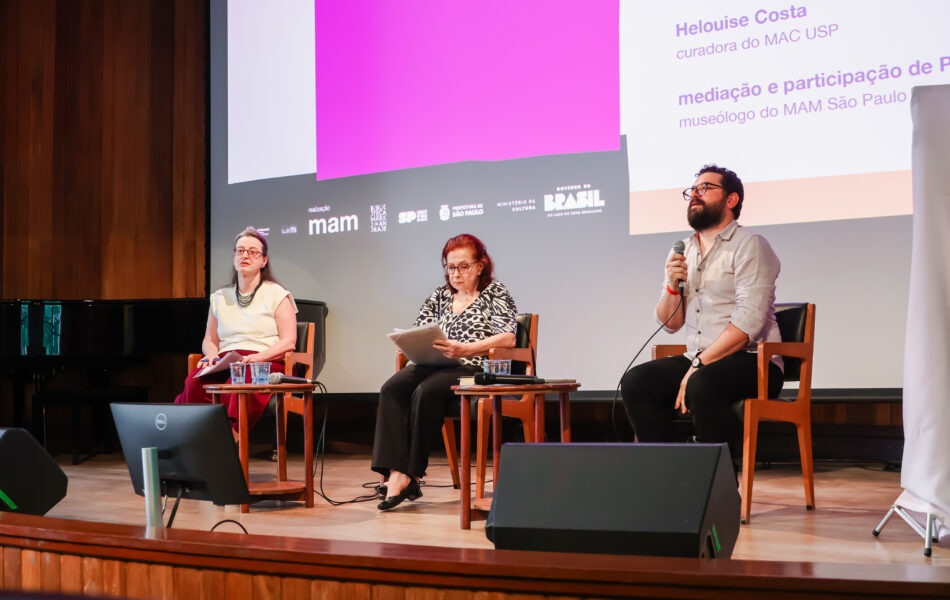 Descrição da imagem: Uma fotografia de um debate em um auditório com fundo de madeira. Três pessoas estão sentadas no palco em cadeiras escuras, separadas por pequenas mesas redondas. O telão ao fundo exibe uma composição gráfica em branco, cinza e roxo com textos sobre curadoria e mediação. À esquerda, uma mulher de blusa clara e saia bordô. No centro, uma mulher de blusa estampada em preto e branco está lendo anotações. À direita, um homem barbudo com óculos e camisa clara está falando ao microfone. Fim da descrição da imagem.
