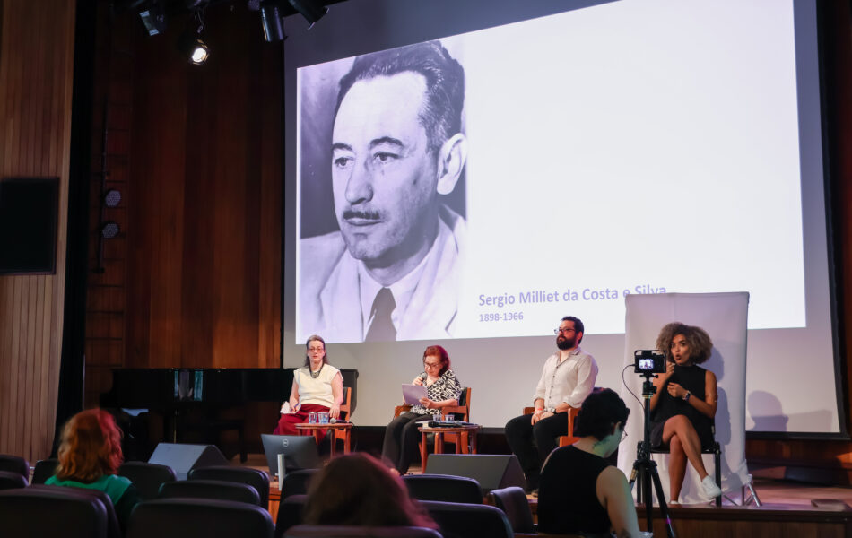 Descrição da imagem: Uma fotografia de um debate em um auditório com fundo de madeira. O telão ao fundo exibe um retrato em preto e branco de Sérgio Milliet da Costa e Silva e seu nome e datas (1898-1966). No palco, três debatedores e uma intérprete de Libras estão sentados. À esquerda, uma mulher de blusa clara e saia bordô. No centro, uma mulher de blusa estampada em preto e branco está lendo anotações. À direita, um homem barbudo com óculos e camisa clara. Uma intérprete de Libras sentada na extrema direita está sendo filmada por uma câmera. O público em primeiro plano é visto de costas. Fim da descrição da imagem.