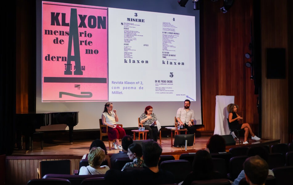 Descrição da imagem: Uma fotografia de um debate em um auditório com fundo de madeira e um grande telão. O telão exibe em fundo branco o texto "A instituição do moderno nos anos 1930 e 1940" em fonte escura. Quatro debatedores e uma intérprete de Libras estão sentados no palco. Um homem de camisa verde e uma mulher de camisa branca estão à esquerda. No centro, um homem com camisa clara está falando ao microfone. Uma mulher de blusa verde escura está sentada à sua direita. Na extrema direita, uma mulher de roupa preta sentada em uma cadeira mais alta está gesticulando em Libras. A plateia no primeiro plano está vista de costas. Fim da descrição da imagem.