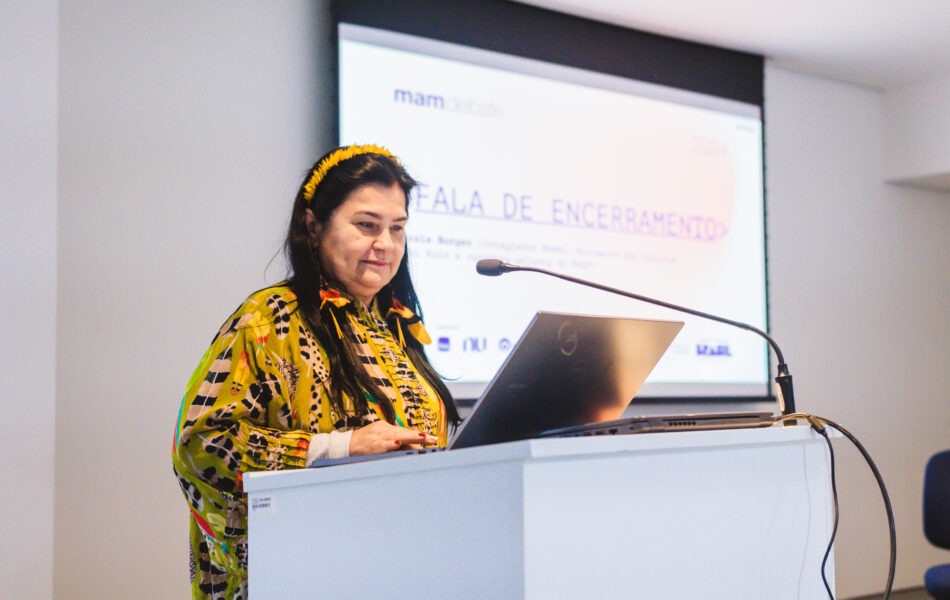 Descrição da imagem: Um close-up de uma mulher fazendo uma fala de encerramento em um debate. A mulher, de cabelo escuro enfeitado com pequenas penas amarelas na cabeça, veste uma blusa estampada em tons de amarelo e verde-oliva. Ela está inclinada sobre um notebook prateado aberto, no púlpito branco, com um microfone de haste posicionado à sua frente. O telão projetado ao fundo exibe o texto "FALA DE ENCERRAMENTO" em letras grandes. A iluminação é forte e artificial. Fim da descrição da imagem.
