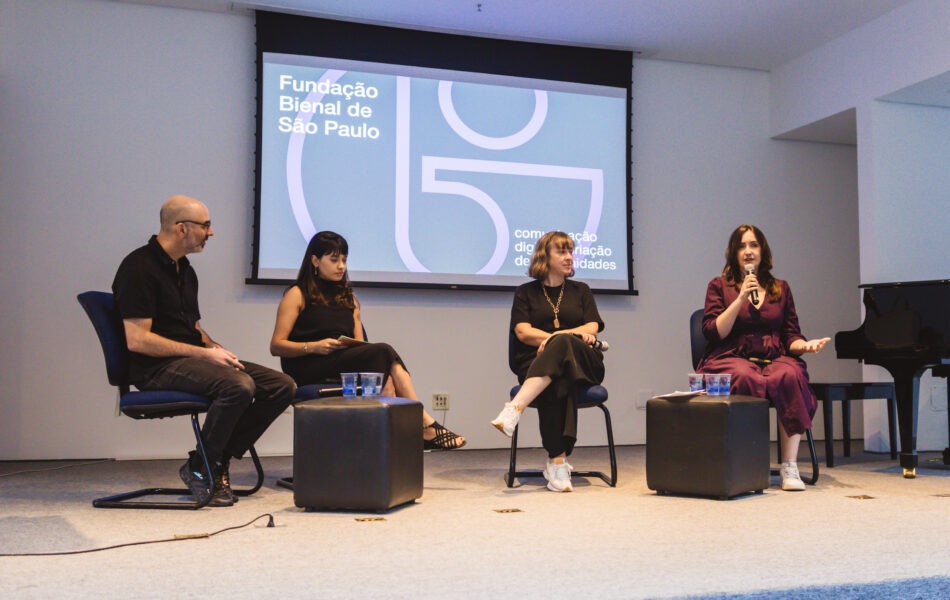 Descrição da imagem: Uma fotografia de uma mesa de debate com quatro participantes sentados em cadeiras azuis, em um palco de paredes brancas. O telão ao fundo exibe o logo da "Fundação Bienal de São Paulo" com o texto "comunicação digital e ativação de comunidades". Da esquerda para a direita: um homem branco e calvo usa camisa preta e calça jeans; uma mulher morena de cabelo liso com franja usa blusa preta; uma mulher branca com cabelo liso e loiro escuro usa camisa preta e calça preta; e uma mulher branca de cabelo liso ruivo escuro usa vestido roxo, falando ao microfone. Eles estão sentados em torno de pequenos pufes quadrados e escuros. Um piano de cauda preto está visível no canto direito. Fim da descrição da imagem.
