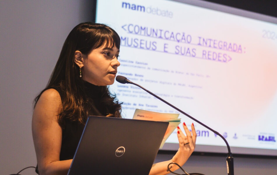 Descrição da imagem: Um close-up de uma mulher no púlpito, apresentando em um evento. A mulher, de cabelo escuro e liso, com franja, veste uma blusa preta sem mangas e gesticula com a mão direita, com unhas vermelhas, enquanto fala ao microfone. Em sua frente, há um notebook preto aberto sobre o púlpito branco. O telão ao fundo exibe o texto "COMUNICAÇÃO INTEGRADA: MUSEUS E SUAS REDES" em azul e roxo. A iluminação é forte e artificial, vinda do alto. Fim da descrição da imagem.