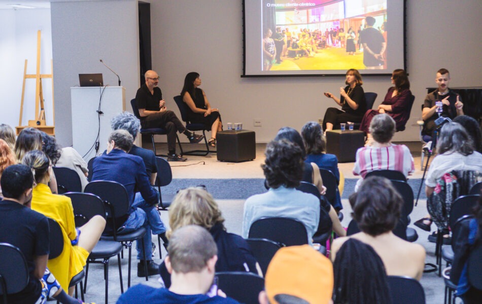 Descrição da imagem: Uma fotografia de plano aberto, capturada de trás da plateia, em um auditório com paredes brancas e teto com luzes fluorescentes. O foco está na plateia, sentada em cadeiras pretas e cinzas. Ao fundo, quatro participantes estão sentados em um palco elevado e escuro. O telão projeta uma imagem colorida de um evento ou performance artística. A iluminação é de teto, com luzes fluorescentes, e a cena tem um aspecto de luz fria. Fim da descrição da imagem.
