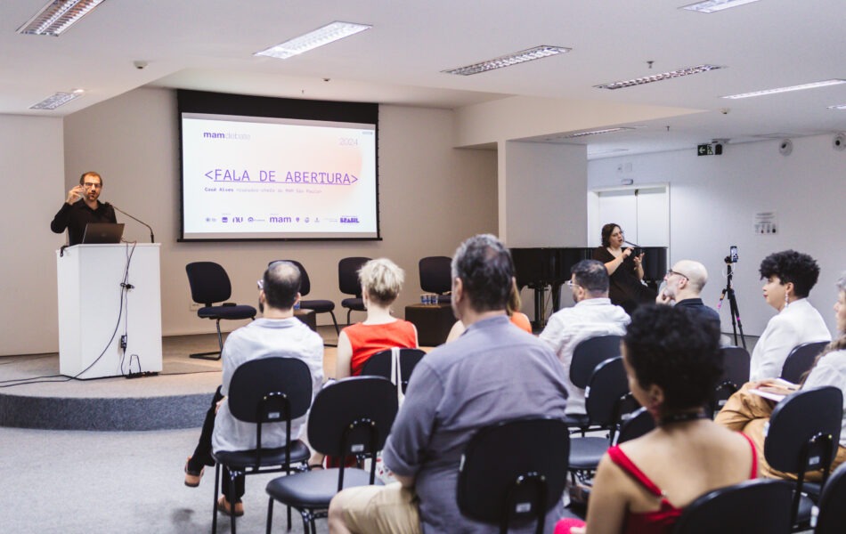 Descrição da imagem: Uma fotografia de um homem no púlpito, fazendo uma fala de abertura em um debate, em um auditório com paredes brancas. O homem, de barba e camisa preta, está falando ao microfone no púlpito branco. O telão ao fundo exibe o texto "mam debate" e "FALA DE ABERTURA". A plateia está sentada em cadeiras pretas, vista de costas. À direita, uma mulher de blusa preta realiza a tradução em Libras. Um piano de cauda preto está visível ao fundo. Fim da descrição da imagem.