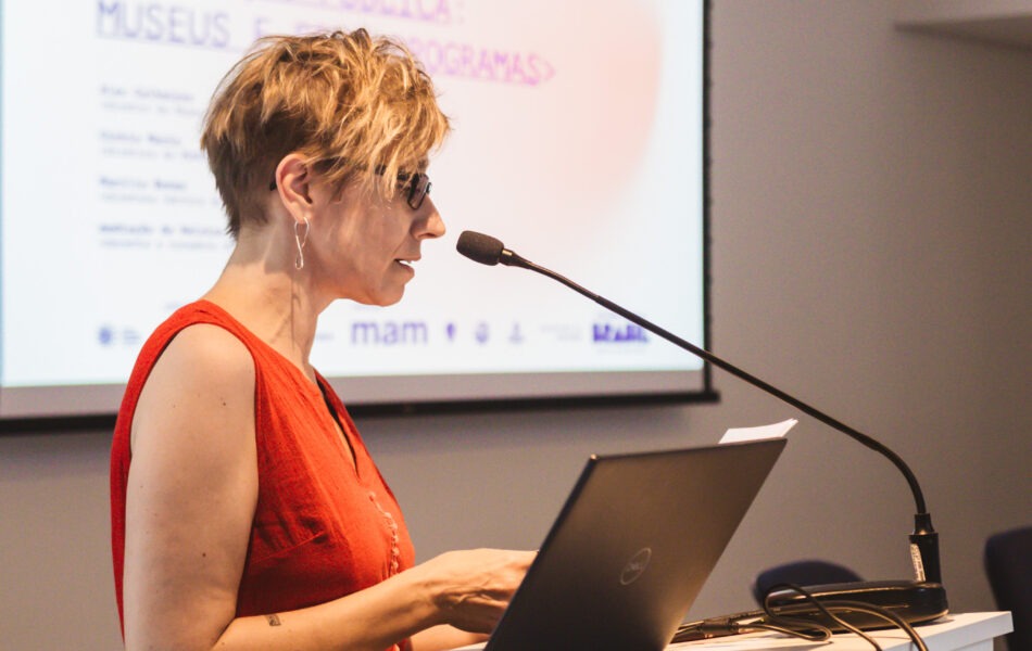 Descrição da imagem: Um close-up de uma mulher loira de cabelo curto no púlpito, apresentando. A mulher, de óculos e vestido vermelho sem mangas, está digitando em um notebook preto no púlpito branco, com o microfone posicionado à sua frente. O telão ao fundo exibe o texto "mam debate" e "PROJEÇÃO PÚBLICA: MUSEUS E PROGRAMAS" em branco e vermelho. A iluminação é forte e artificial, destacando o perfil da oradora. Fim da descrição da imagem.