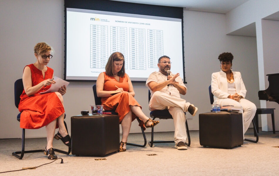 Descrição da imagem: Uma fotografia de uma mesa de debate com quatro participantes sentados em cadeiras azuis escuras, em um palco de paredes brancas. O telão ao fundo exibe uma tabela com o título "NÚMERO DE VISITANTES (1945-2024)". Da esquerda para a direita: uma mulher loira e branca usa vestido vermelho; uma mulher branca e loira escuro usa vestido laranja; um homem branco, barbudo e de óculos usa roupa branca e está gesticulando ao microfone; e uma mulher negra de cabelo crespo usa roupa branca. Eles estão sentados entre pequenos pufes quadrados e escuros. Um piano de cauda preto está visível no canto direito. Fim da descrição da imagem.