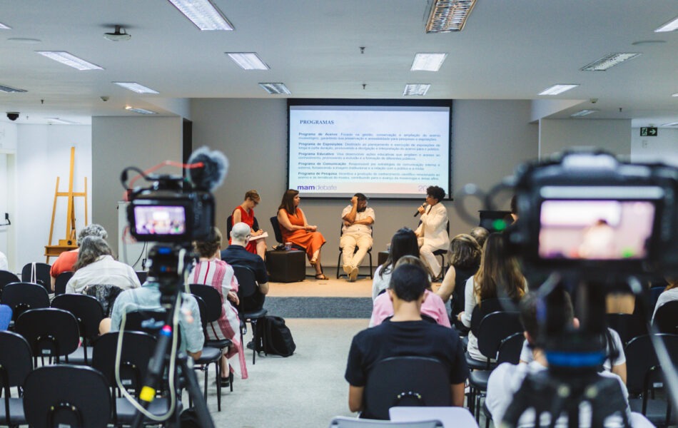 Descrição da imagem: Uma fotografia em plano aberto de uma mesa de debate, vista de trás da plateia, focada em duas câmeras de vídeo em tripés no primeiro plano. Quatro participantes estão sentados no palco, em cadeiras pretas. O telão ao fundo exibe o título "PROGRAMAS" e uma lista de tópicos em letras pretas sobre fundo branco, com o logo "mam" abaixo. A plateia está sentada em cadeiras pretas, e a iluminação é de teto, com luzes fluorescentes. Fim da descrição da imagem.