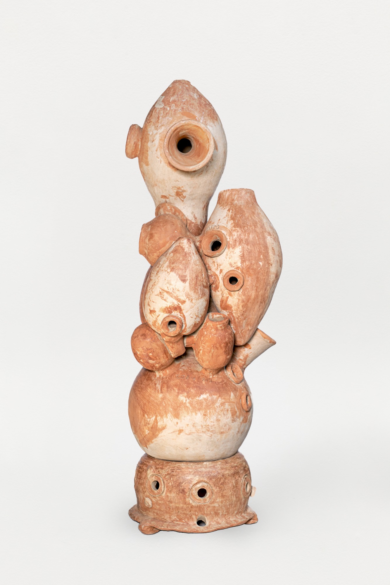 Descrição da imagem: Escultura vertical em cerâmica, com tons terrosos, composta por várias formas sobrepostas com orifícios circulares, fundos, e alguns com bordas projetadas para fora. No topo, uma forma ovalada e alongada, voltada para cima, apresenta um orifício central e outro menor na lateral, ambos circulares e com bordas projetadas para fora. Abaixo, um aglomerado de formas ovaladas e arredondadas, de tamanhos variados, exibe orifícios de diferentes dimensões, também circulares e de bordas projetadas para fora. Próximo à base, uma grande forma arredondada tem um pequeno bocal alongado projetado para a direita. O conjunto está apoiado sobre uma forma baixa e arredondada, com três pequenos orifícios ao redor, também circulares. Fim da descrição da imagem.
