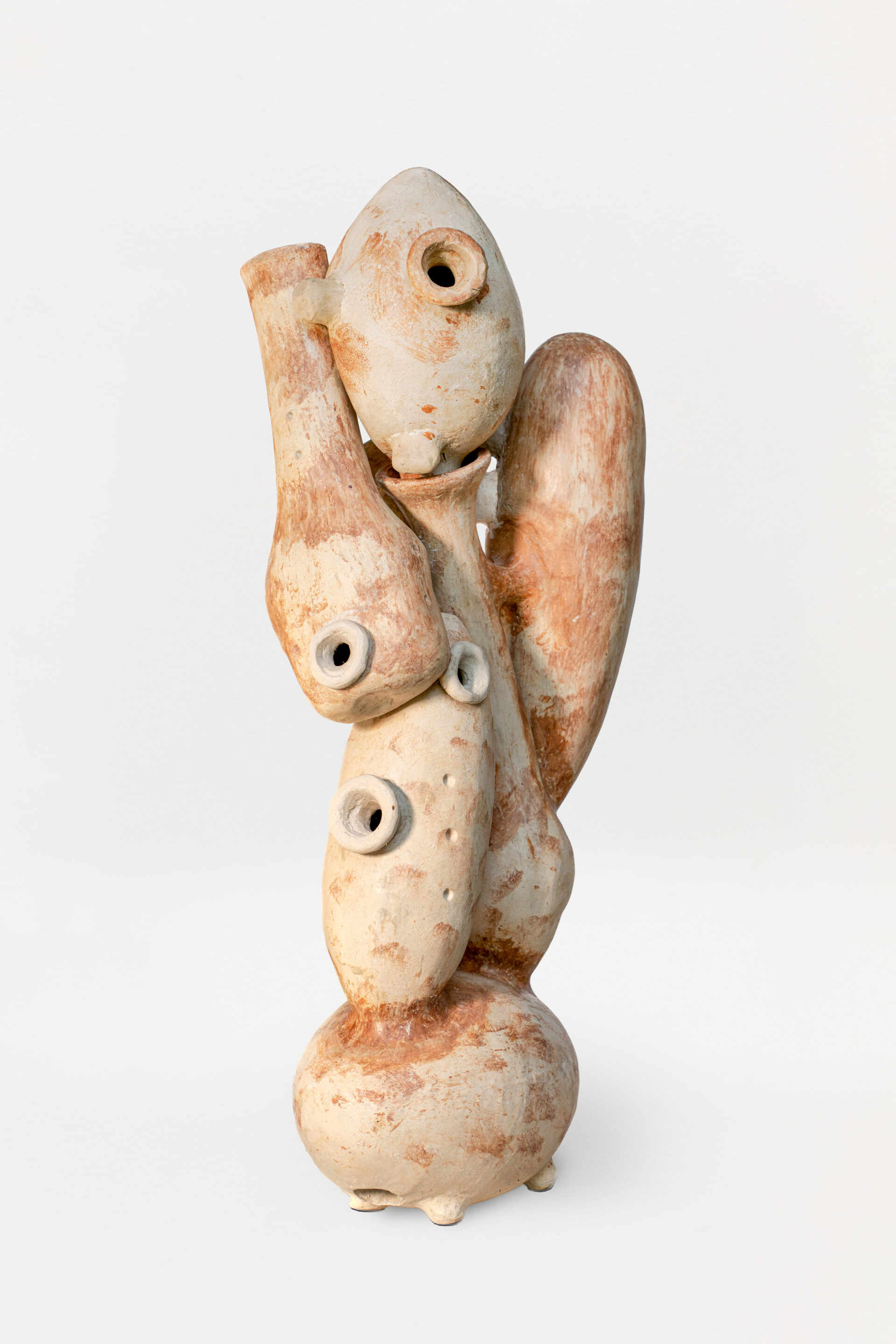 Descrição da imagem: Escultura vertical em cerâmica, com tons terrosos, composta por várias formas sobrepostas com orifícios circulares, fundos, e alguns com bordas projetadas para fora. Na parte superior, há uma forma ovalada com um pequeno orifício central e circular com a borda projetada para fora. À esquerda, uma forma tubular se projeta para cima, com um orifício circular na parte inferior também com a borda projetada para fora. Abaixo, há uma forma alongada e vertical, com orifício no centro e outro circular na lateral, ambos circulares e de bordas projetadas para fora. No centro, uma forma longa e lisa está levemente inclinada na diagonal, e à direita, outra forma longa, lisa e larga. A base da escultura é arredondada na horizontal e apoiada sobre três pezinhos arredondados. Fim da descrição da imagem.
