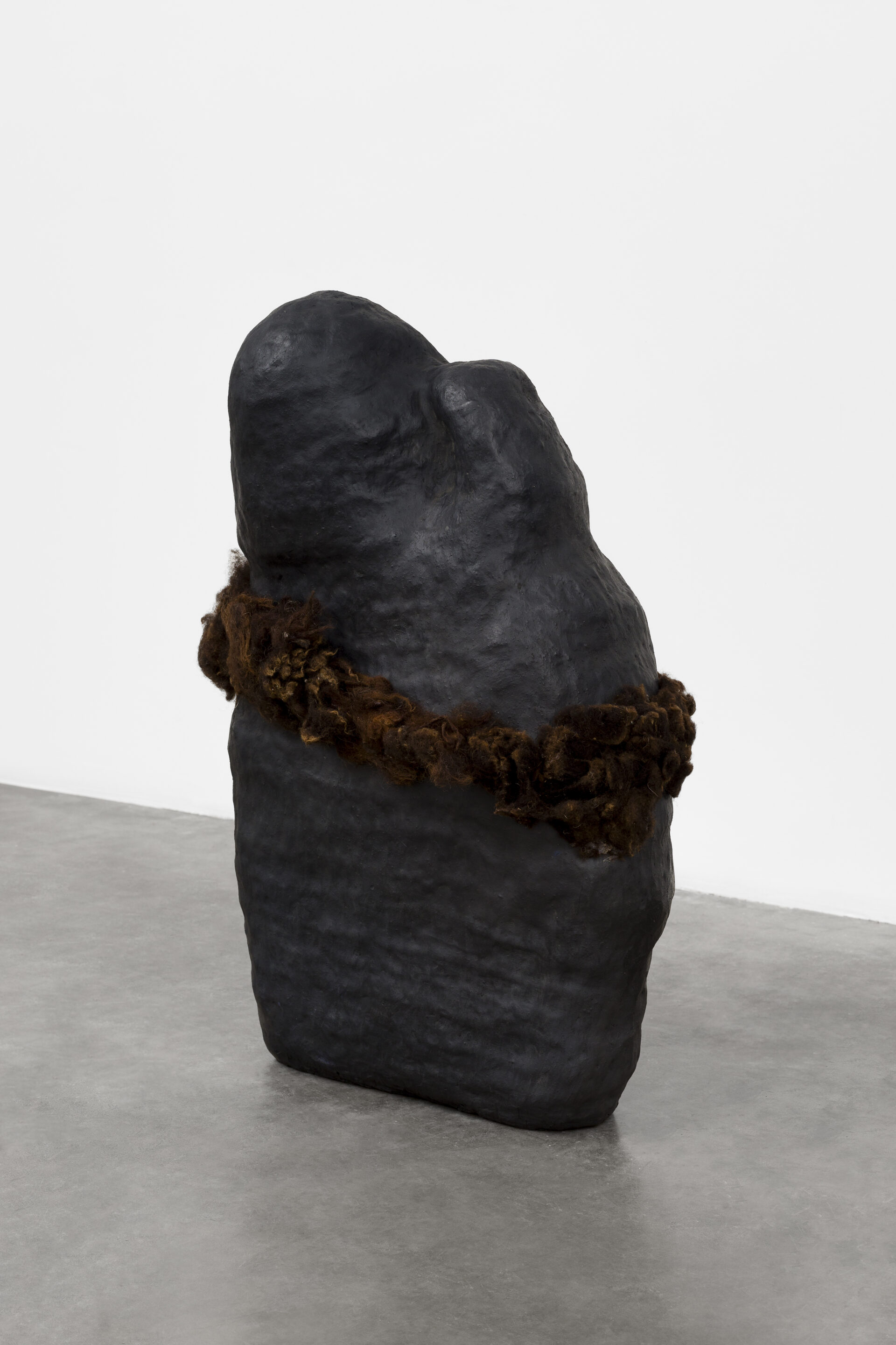 Descrição da imagem: Escultura de cerâmica em formato ovalado, vertical, larga e irregular, semelhante a uma grande pedra, de tamanho médio, posicionada sobre chão claro. A obra é preta-acinzentada, de textura opaca. Ao redor da parte central, há uma faixa volumosa e densa de fios grossos e entrelaçados em tons de marrom.  Fim da descrição da imagem.
