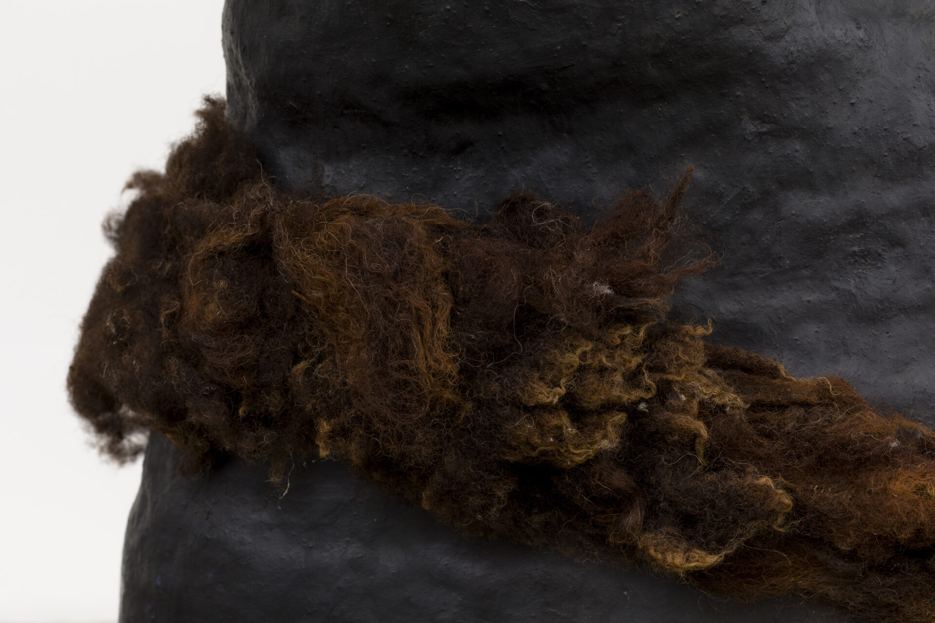 Descrição da imagem: Fotografia horizontal. Vista de perto, a parte central da escultura de cerâmica em formato ovalado, vertical, larga e irregular, com a faixa volumosa e densa de fios grossos e entrelaçados em tons de marrom. Fim da descrição da imagem.
