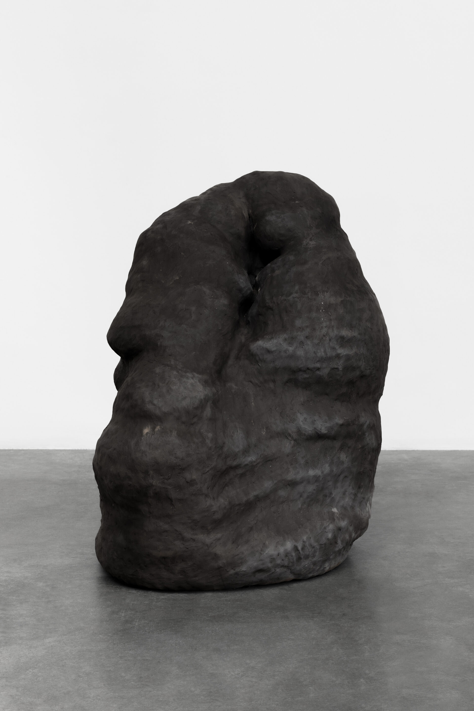 Descrição da imagem: Escultura de cerâmica em formato ovalado, vertical, larga e irregular, semelhante a uma grande pedra, de tamanho médio, posicionada sobre chão claro. A obra é preta-acinzentada, de textura opaca e levemente ondulada, com saliências sutis e uma cavidade na parte superior. Fim da descrição da imagem.
