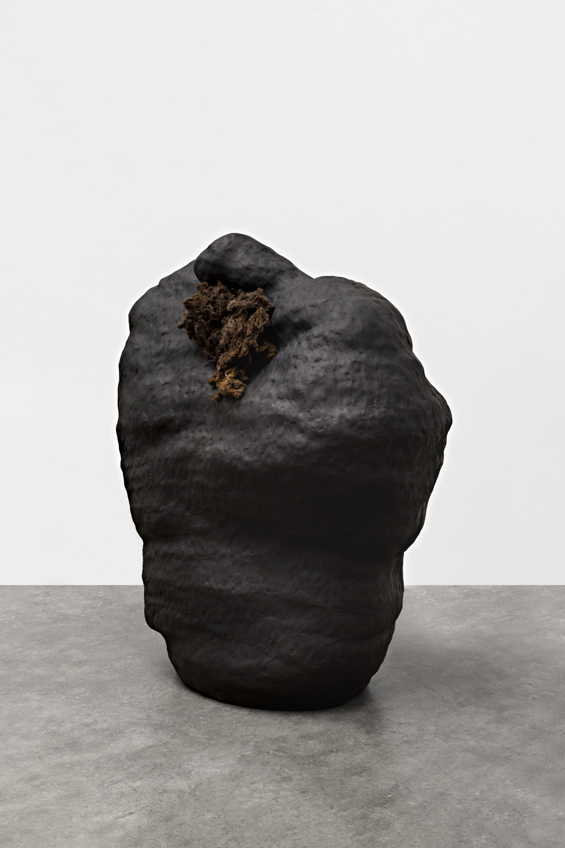 Descrição da imagem: Escultura de cerâmica em formato ovalado, vertical, larga e irregular, semelhante a uma grande pedra, de tamanho médio, posicionada sobre chão claro. A obra é preta-acinzentada, de textura opaca, levemente ondulada e com saliências sutis. Na parte superior, ao centro, inserida em uma cavidade, há um emaranhado de lã marrom.  Fim da descrição da imagem.
