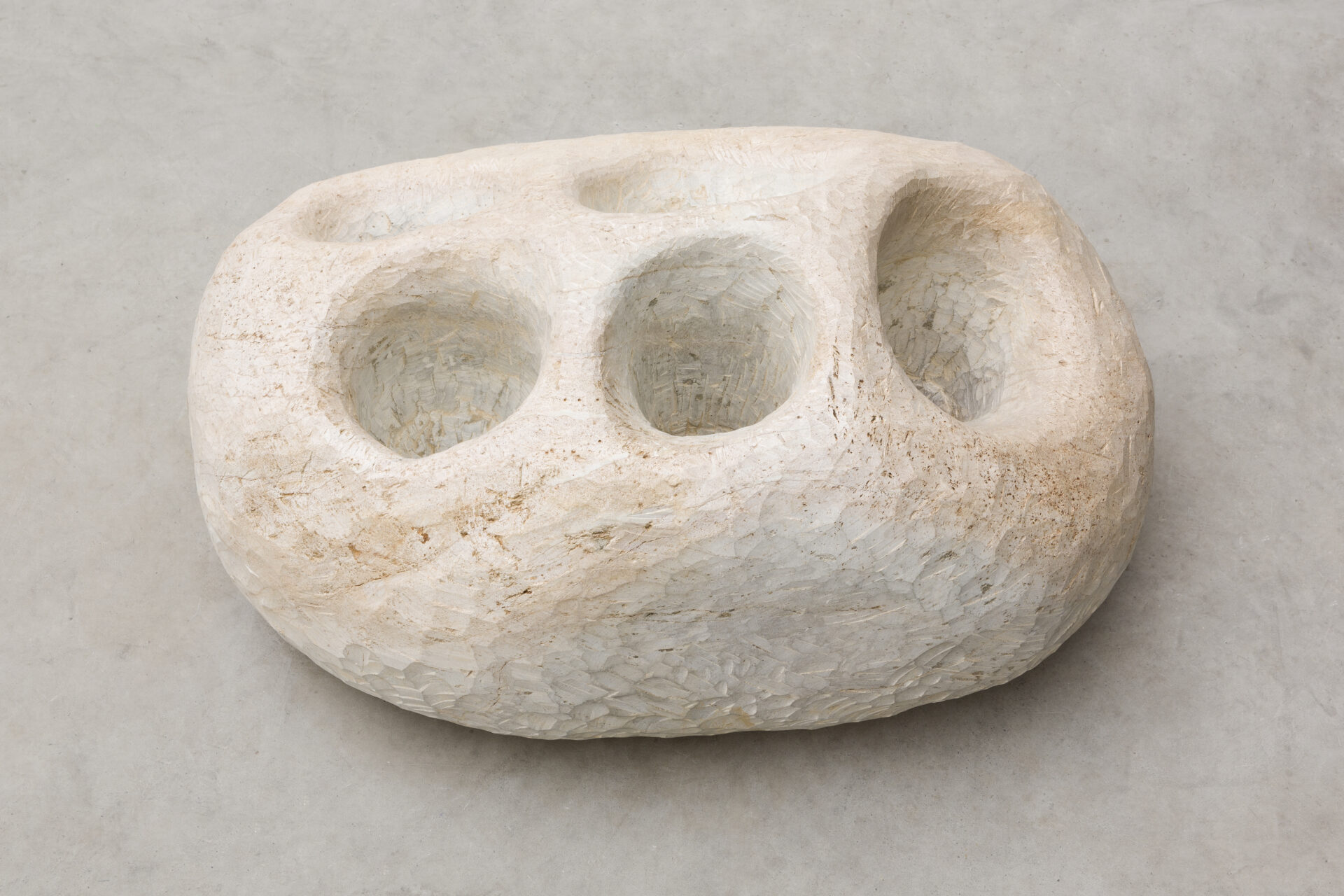 Descrição da imagem: Escultura de pedra-sabão posicionada sobre chão claro. A superfície é em tom de branco gelo, com pequenas manchas amarronzadas. O formato é ovalado e irregular, e na parte superior há três cavidades arredondadas de profundidades diferentes. Em uma das laterais, outras duas cavidades também arredondadas. Fim da descrição da imagem.
