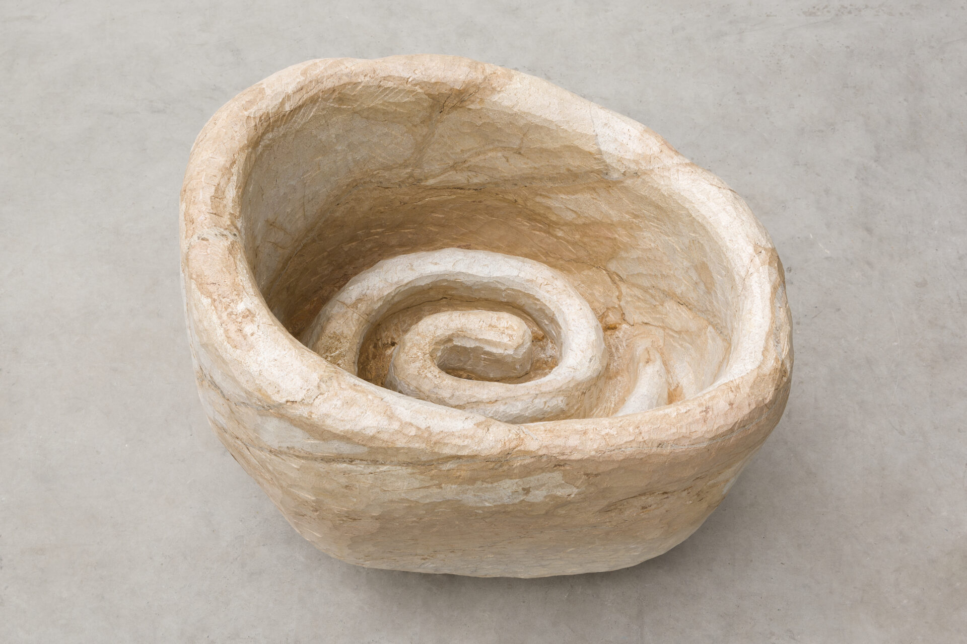 Descrição da imagem: Escultura de pedra-sabão posicionada sobre chão claro. A superfície é em tom de branco gelo, com manchas amarronzadas, e nas paredes internas há poucas fissuras. O formato é circular e irregular, com interior côncavo de bordas espessas, sendo um dos lados mais alto que o outro. O centro tem formato espiralado em alto-relevo. Fim da descrição da imagem.
