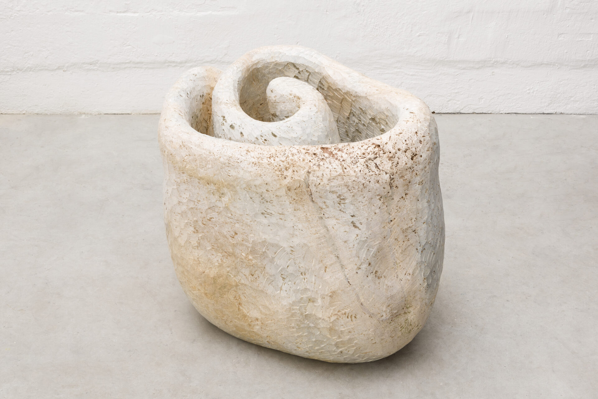 Descrição da imagem: Escultura de pedra-sabão posicionada sobre chão claro. A superfície é em tom de branco gelo, com manchas e pequenos pontos amarronzados. O formato é espiralado e irregular, com interior côncavo de média profundidade e bordas espessas.  Fim da descrição da imagem.