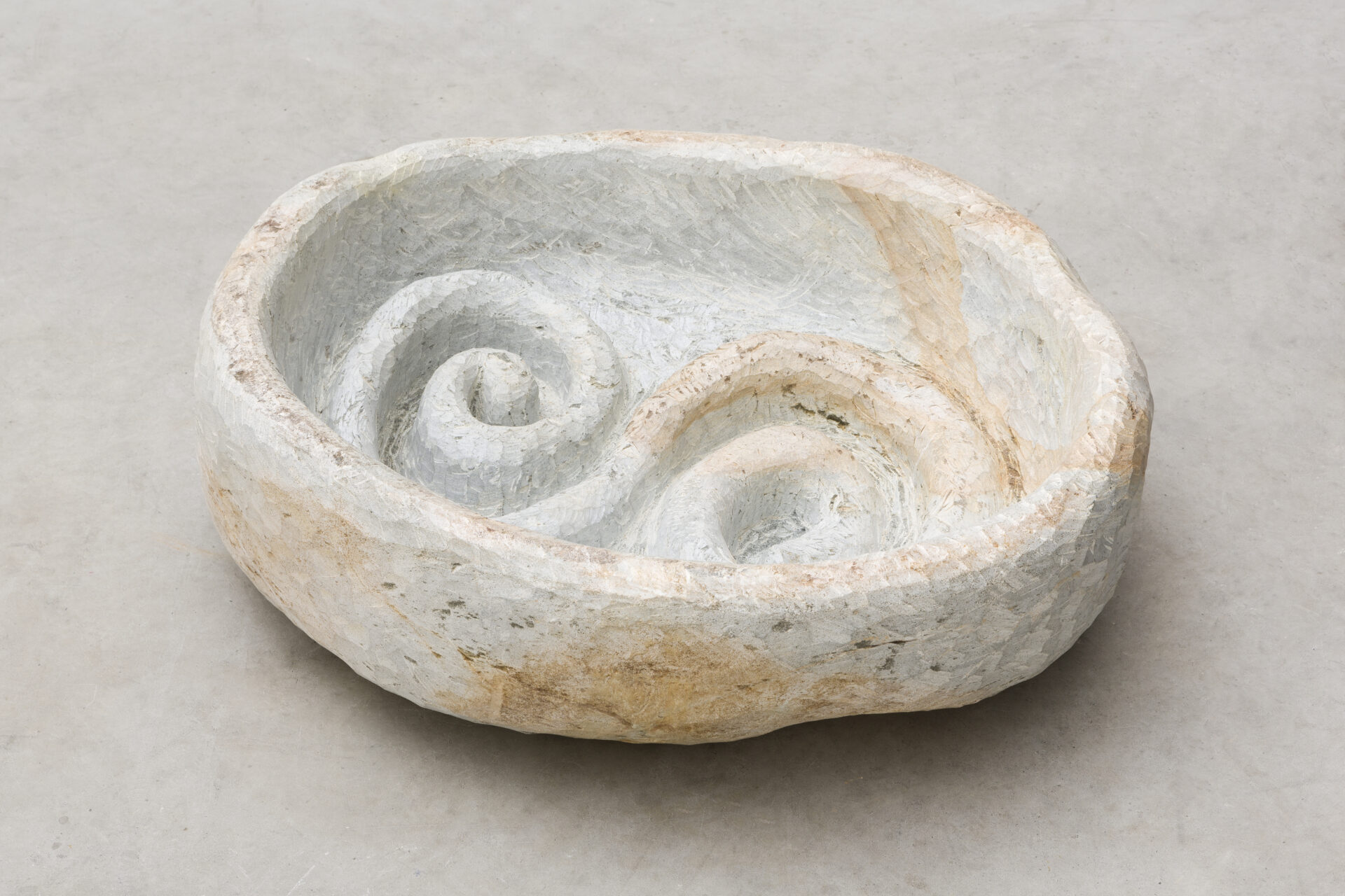 Descrição da imagem: Escultura de pedra-sabão posicionada sobre chão claro. A superfície é em tom de branco gelo, com manchas amarronzadas. O formato é circular e irregular, com interior côncavo de bordas espessas. No centro, há duas espirais contínuas.  Fim da descrição da imagem.
