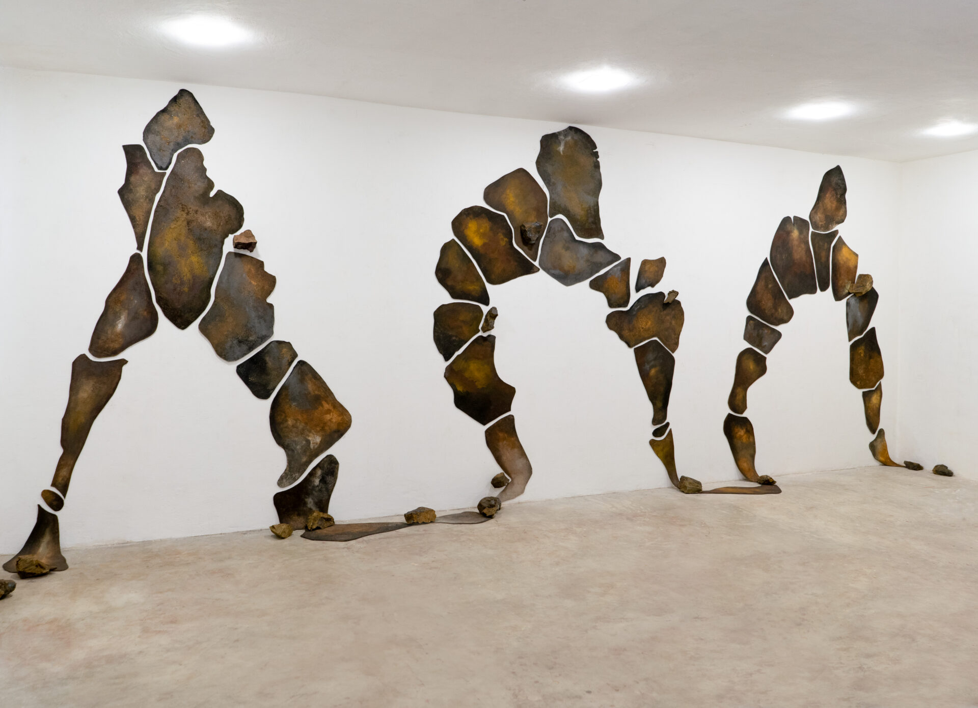 Descrição da imagem: Instalação composta por quarenta placas irregulares revestidas por lona crua tingida com pigmentos minerais em tons de marrom-escuro e nuances amareladas.
As placas, de textura terrosa e aparência densa, estão fixadas em uma parede branca e organizadas em três conjuntos verticais que formam arcos irregulares.
Algumas se estendem até o chão de concreto, onde outras placas estão apoiadas junto à base.
Nas laterais, pedras nas mesmas tonalidades estão posicionadas próximas às placas inferiores. Fim da descrição da imagem.
