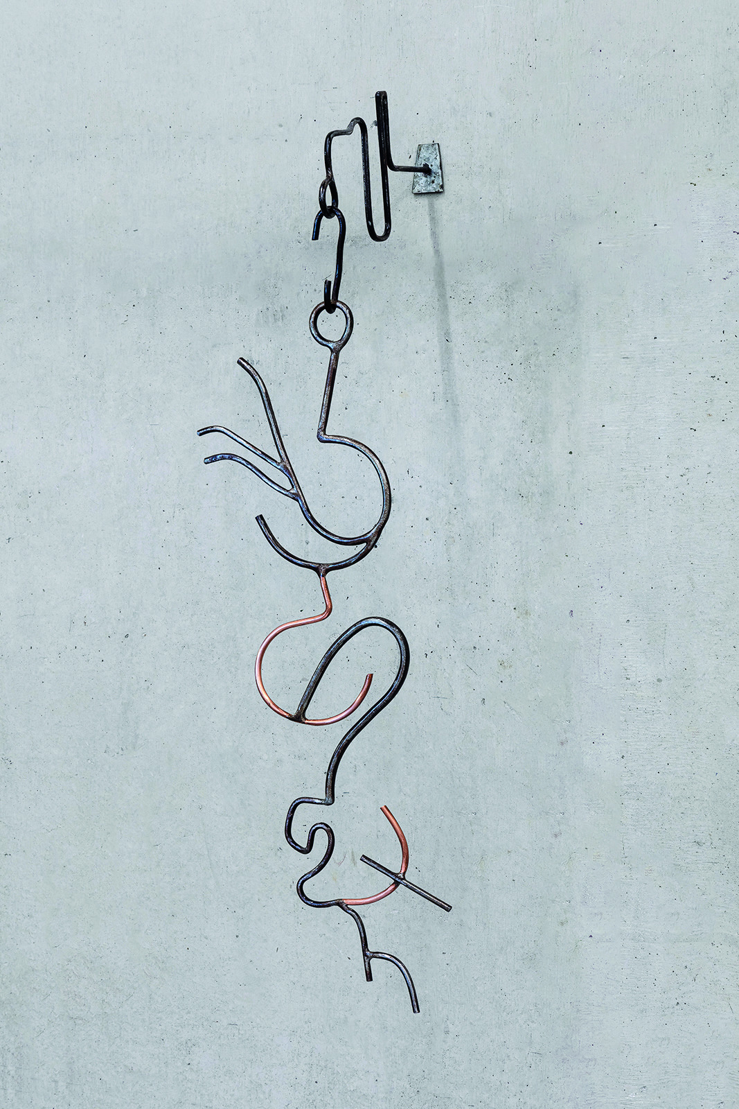 Descrição da imagem: Escultura de ferro e cobre retorcidos, suspensa por um suporte metálico fixado em uma parede de concreto. A peça é composta por aproximadamente sete hastes metálicas finas e curvas, que formam linhas sinuosas entrelaçadas verticalmente. As hastes, em tons escuros com partes em cobre avermelhado, criam formas abstratas. Fim da descrição da imagem.
