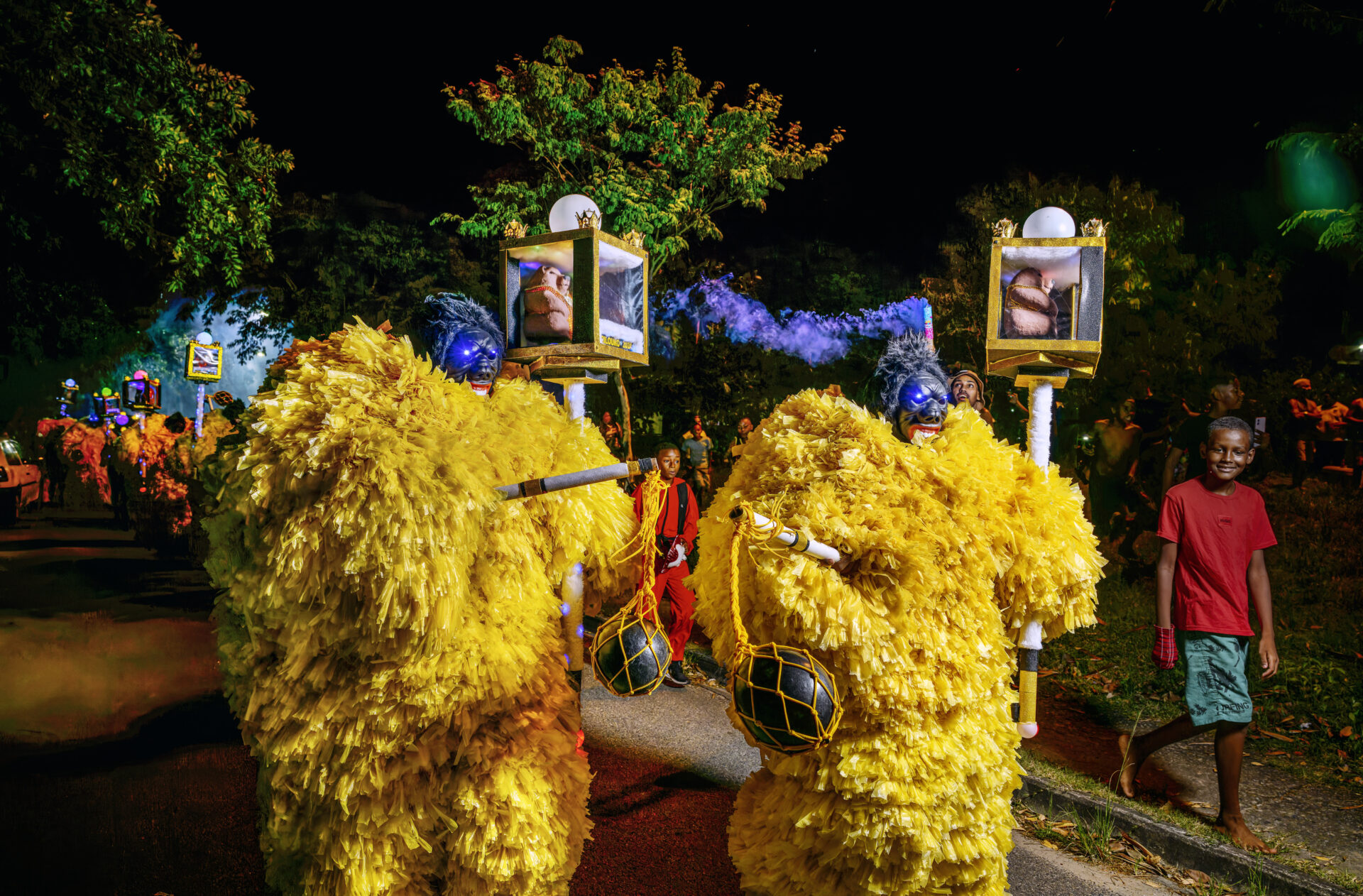 Descrição da imagem: Fotografia noturna, em formato horizontal, de um grupo com aproximadamente vinte Guriloukos. Da frente para o fundo, eles estão enfileirados em uma rua asfaltada. Diante do grupo, dois Guriloukos usam máscaras azul-escuras, com cabelos curtos da mesma cor; seguram o bastão na mão direita e, na esquerda, uma haste branca que sustenta uma caixa quadrada translúcida, com molduras douradas. Dentro, um urso marrom está sentado. Acima da caixa, há um pequeno globo branco.  À direita, um menino negro, com o olhar voltado para a frente, sorri. Tem cabelos raspados, sobrancelhas e lábios finos. Usa camiseta vermelha, bermuda verde e uma luva vermelha na mão direita. Ao redor, outras pessoas observam. Ao fundo e na lateral direita, há árvores altas com copas esverdeadas. Fim da descrição da imagem.
