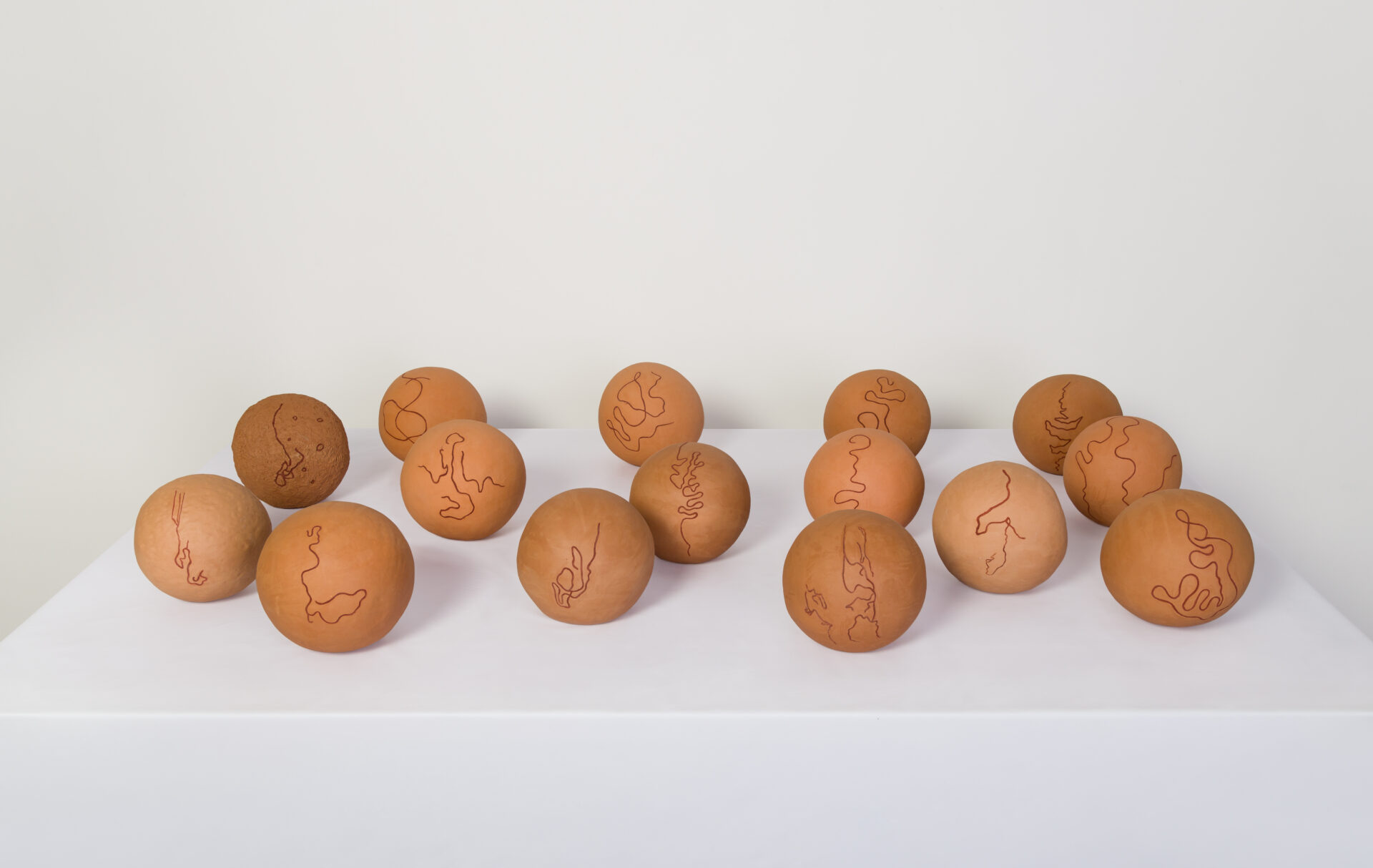 Descrição da imagem: Conjunto de quinze esferas de cerâmica dispostas sobre uma superfície branca. As peças têm tamanhos semelhantes e tons que variam do marrom-claro ao alaranjado. Cada esfera apresenta marcações sinuosas, gravadas na superfície com pigmento escuro feito com óxido de ferro. Os traços finos formam desenhos abstratos e únicos em cada peça. Fim da descrição da imagem.
