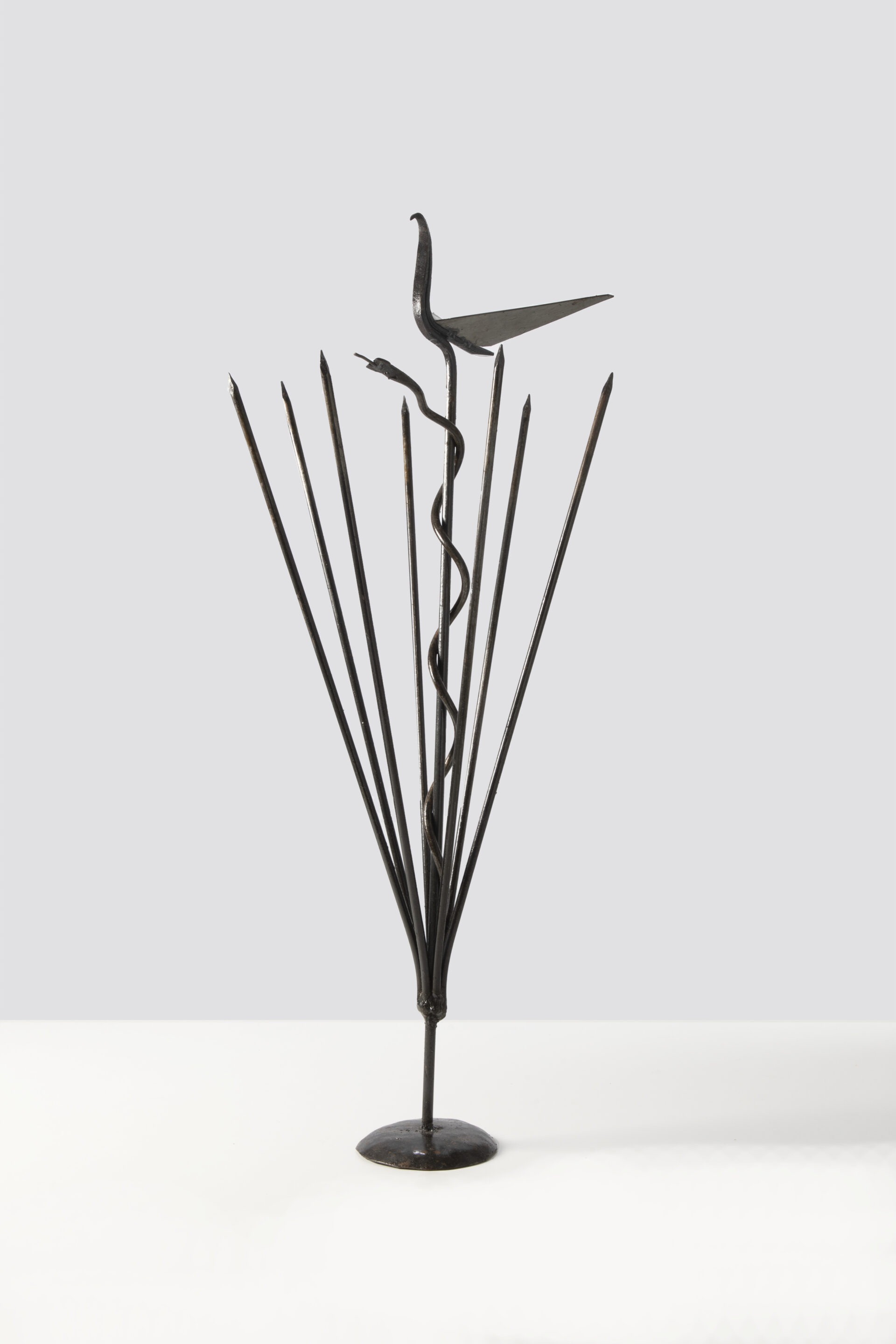 Descrição da imagem: Ferramenta de Agué. A peça é sustentada por uma base circular, apoiada sobre chão claro.
Estrutura metálica vertical, escura, composta por hastes de ferro.
De uma haste central reta, erguem-se, na parte inferior, sete hastes levemente posicionadas em diagonal, sendo quatro à esquerda e três à direita, todas com pontas finas.
Da base da haste central, uma haste representando uma cobra está entrelaçada em espiral até a parte superior, onde há uma ave de ferro, de corpo fino e ereto, com asas em formato triangular abertas. Fim da descrição da imagem.
