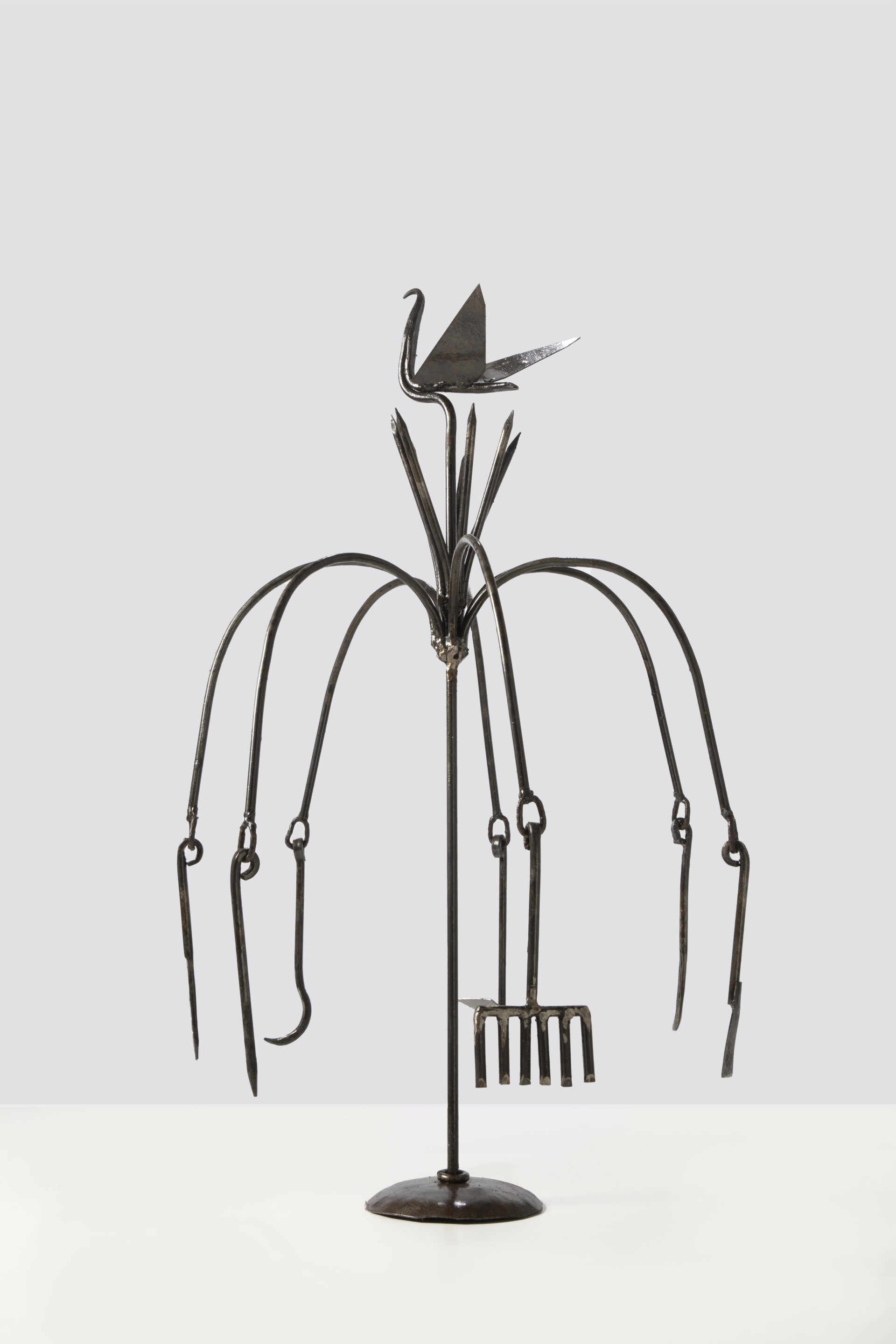 Descrição da imagem: Ferramenta de Ogum Onirê. A peça é sustentada por uma base circular, apoiada sobre chão claro.
Estrutura metálica vertical, escura, composta por hastes de ferro. Ao redor da haste central reta, erguem-se, no alto, sete hastes curvadas para fora e para baixo. Nas extremidades, pendem diferentes formas metálicas alongadas: ganchos, lâminas estreitas e um rastelo de seis dentes.
Acima, projetam-se, ao redor da haste central, seis hastes retas apontadas para o alto. No topo da haste central, há uma ave de ferro, de corpo fino e ereto, com asas em formato triangular abertas.  Fim da descrição da imagem.

