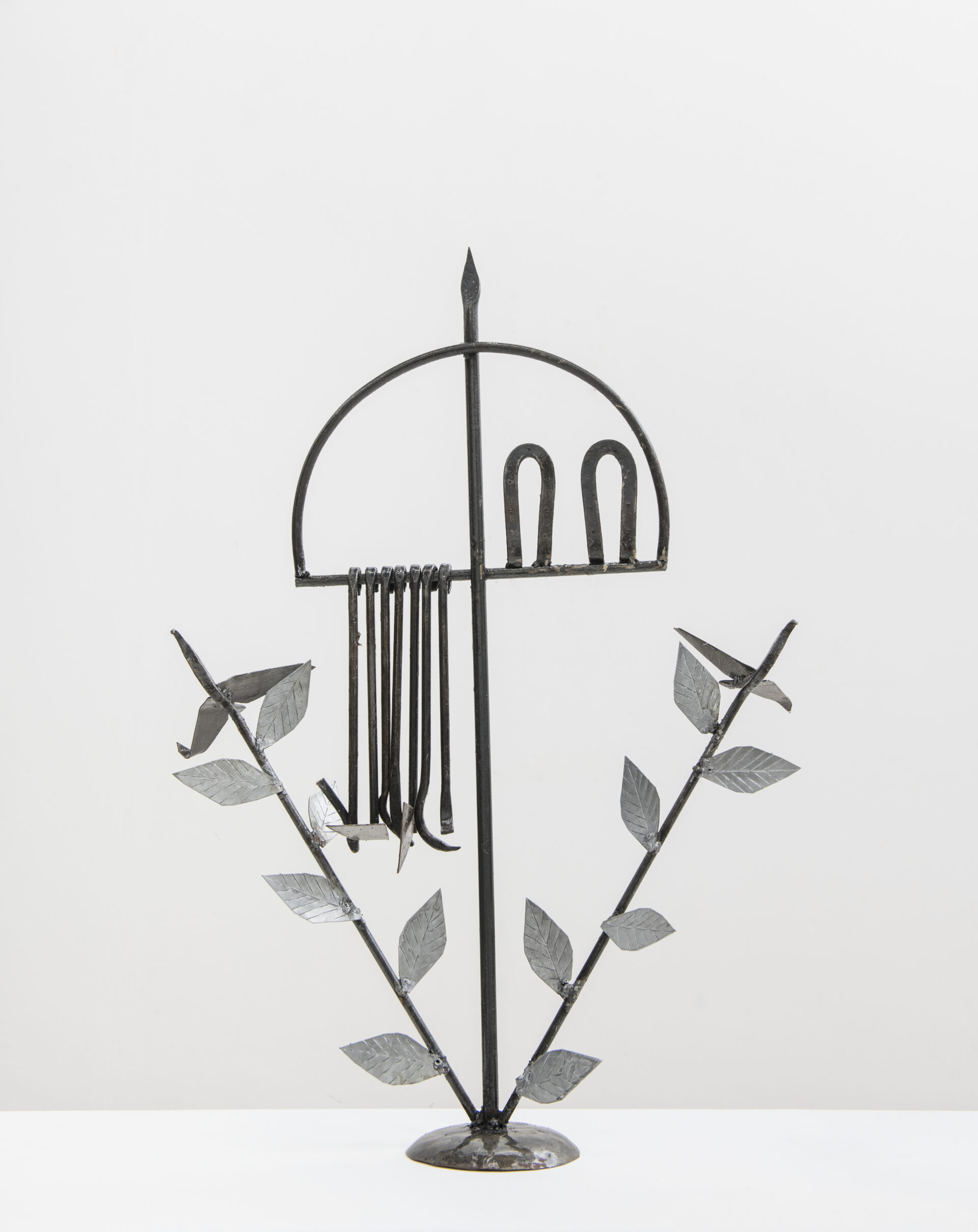 Descrição da imagem: Ferramenta de Oxóssi Odé. A peça é sustentada por uma base circular, apoiada sobre chão claro.
Estrutura metálica vertical, escura, composta por hastes de ferro. No centro, uma haste reta representa uma flecha que atravessa um arco, formando um ofá, ferramenta de caça de Oxóssi. O arco tem formato semicircular e está apoiado horizontalmente na flecha.
Na parte inferior do arco, do lado esquerdo, pendem cinco hastes com pequenas ferramentas agrícolas; do lado direito, duas hastes em formato de arco, voltadas para cima.
Nas laterais da base da haste central, há duas hastes posicionadas em diagonal, com folhas metálicas alongadas e pontudas, de nervura central marcada, assemelhando-se a folhas de cipó, distribuídas ao longo de cada haste e, nas pontas voltadas para fora, aves de ferro de corpo fino e ereto, com asas em formato triangular abertas. Fim da descrição da imagem.
