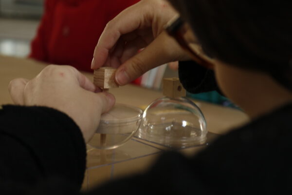 Imagem. Início da descrição de imagem: Fotografia em close-up focada nas mãos de uma criança que participa de uma atividade educativa. As mãos manuseiam pequenas peças de um jogo pedagógico composto por cubos de madeira e cúpulas de acrílico transparente. Em primeiro plano, a criança encaixa um cubo de madeira sobre um pequeno pilar central dentro de uma das cúpulas. Ao lado, há outra cúpula já montada com um cubo no topo. A iluminação é suave, destacando a concentração no trabalho manual. Fim da descrição de imagem.