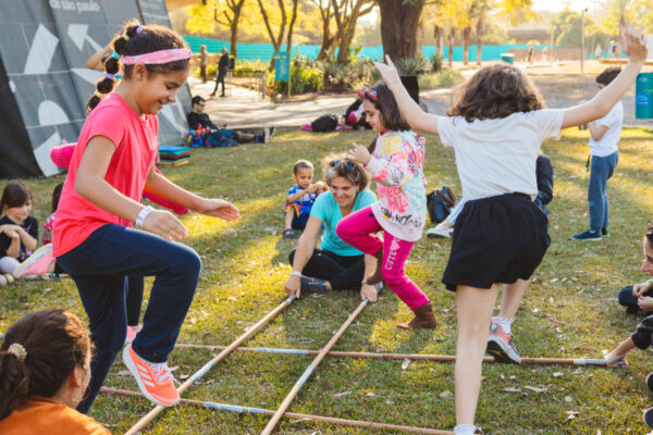 Imagem. Início da descrição de imagem: Fotografia colorida capturada ao ar livre no Jardim de Esculturas do MAM, sob a luz do sol de fim de tarde. Um grupo de crianças participa de uma brincadeira de saltar sobre varas de bambu dispostas em cruz no gramado, as quais são movimentadas por adultos agachados. Em destaque, uma menina à esquerda veste camiseta rosa e calça azul, saltando com um sorriso, enquanto outra criança à direita, de camiseta branca e saia preta, pula com os braços abertos. Ao fundo, vê-se a arquitetura de concreto curva do museu, árvores altas e outras pessoas sentadas na grama. Fim da descrição de imagem.