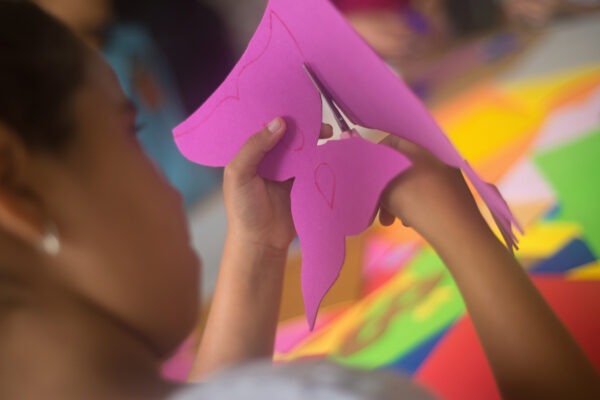 Imagem. Início da descrição de imagem: Fotografia em close-up focada nas mãos de uma criança que utiliza uma tesoura de metal para recortar uma máscara em uma folha de EVA rosa vibrante. O recorte segue o contorno de um desenho feito previamente com caneta vermelha sobre o material. Ao fundo, de forma desfocada, veem-se outras folhas de EVA coloridas sobre uma mesa de ateliê. Fim da descrição de imagem.