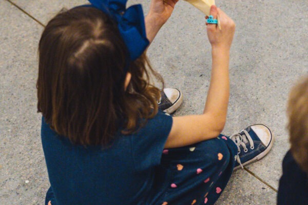 Imagem. Início da descrição de imagem: Uma menina sentada no chão é vista de costas enquanto segura um origami de papel bege em formato de pássaro. Ela usa um vestido azul escuro com estampa de corações coloridos na parte da saia e um grande laço azul no cabelo. Ao seu redor, outras pessoas estão sentadas no chão de granilite claro, sugerindo um momento de mediação cultural ou oficina artística. Fim da descrição de imagem.