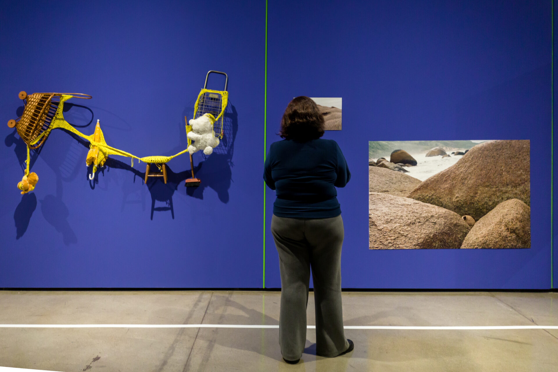 Descrição da imagem:  Fotografia horizontal. Em um ambiente com piso claro marcado por uma linha branca horizontal, uma mulher é vista de costas diante de uma parede azul. Na parede, há uma linha verde vertical. A mulher está posicionada atrás da linha branca e à direita da linha verde.
A mulher tem cabelos lisos e castanhos na altura dos ombros e usa camiseta azul-escura de mangas longas e calça cinza-escura.
À esquerda, fixada à parede, uma instalação conectada por um tecido de crochê amarelo, formada por um pequeno carrinho de bebê composto por um cesto de fibras marrom-escuras; abaixo, uma pelúcia suspensa pelo tecido; ao lado, em direção à direita, um guarda-chuva amarelo, um banquinho de madeira clara, uma pequena vassoura de brinquedo, um ursinho branco visto de costas e um pequeno carrinho metálico de feira. À direita, duas fotografias exibem paisagens com grandes rochas em tons de marrom; a menor está posicionada na parte superior, ao lado da linha verde, e a maior está posicionada à direita, na parte inferior. Fim da descrição da imagem.
