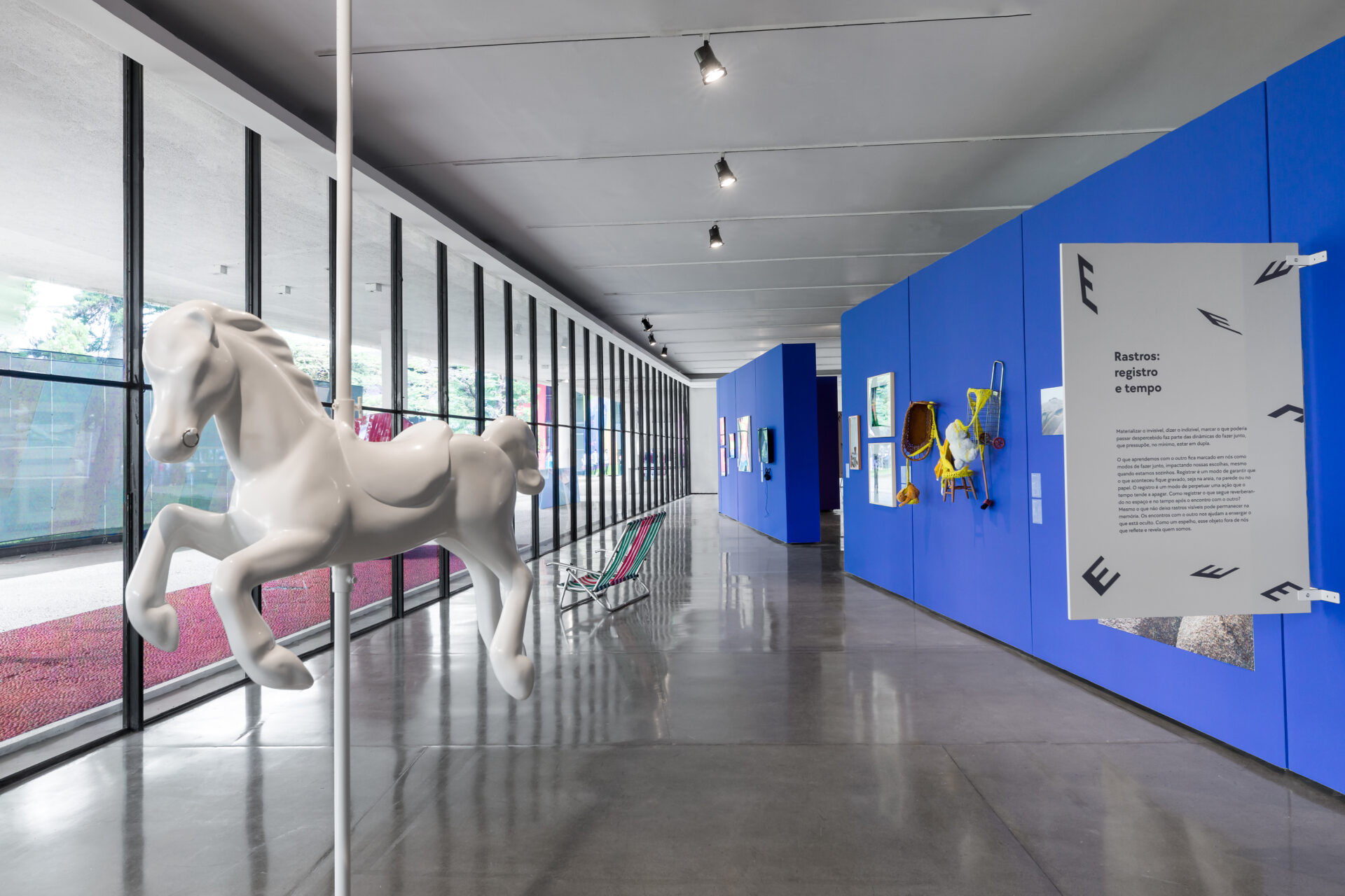 Descrição da imagem:  Fotografia horizontal de um corredor expositivo com teto branco e piso de cimento liso e refletivo. A parede da esquerda é formada por painéis de vidro entremeados por esquadrias metálicas pretas. A da direita é azul e exibe várias obras, como pinturas e instalações. À frente e à esquerda, uma escultura branca de cavalo de carrossel está suspensa por uma haste presa ao teto e ao piso.  Fim da descrição da imagem.
