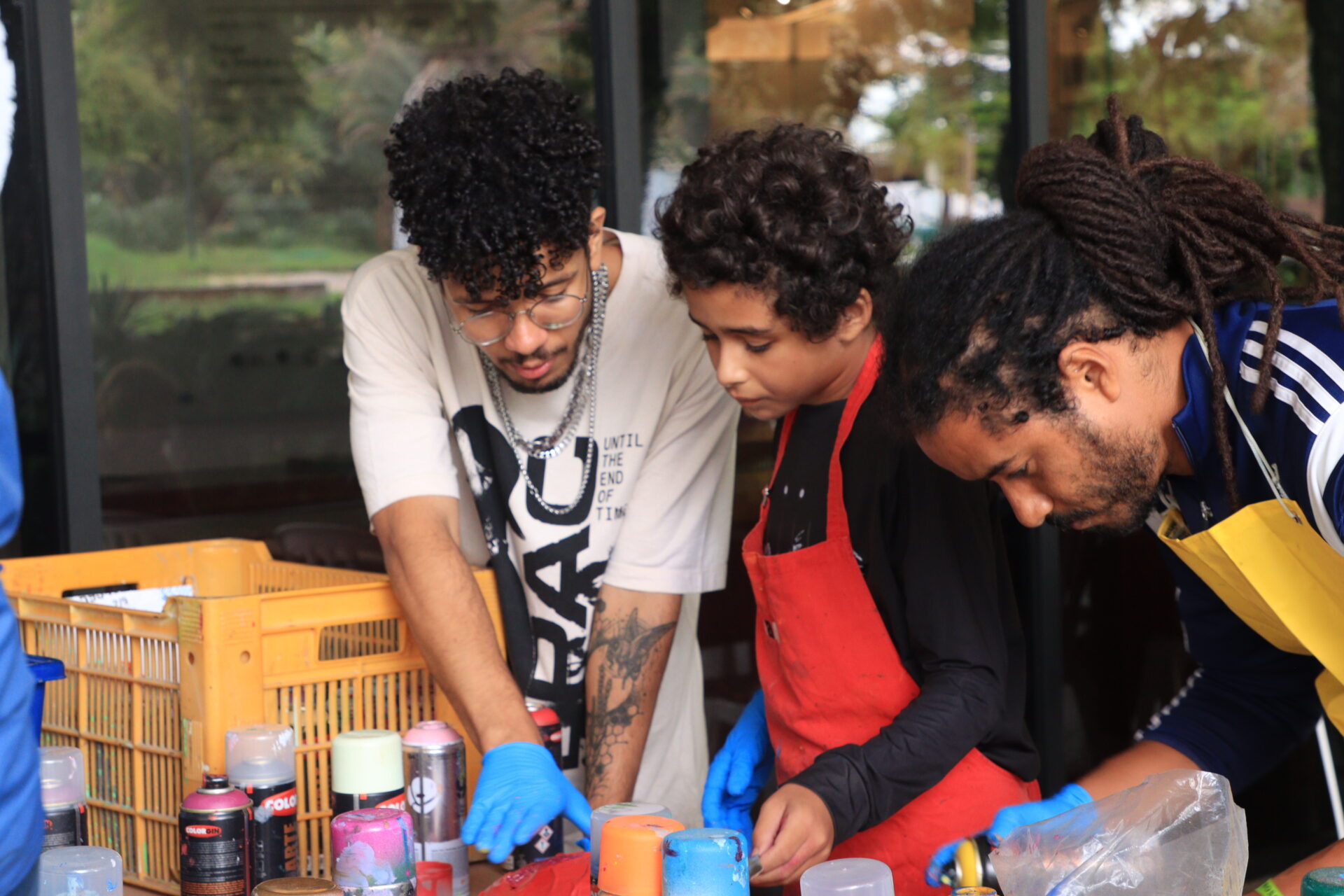 Descrição da imagem: Fotografia horizontal de Daniel Cruz e mais duas pessoas diante de uma mesa com vários sprays de tinta.
À esquerda, Daniel é um homem negro de pele clara, de cabelos pretos e cacheados na altura da testa, com sobrancelhas médias, nariz arredondado na ponta, bigode e barba pretos. Usa camiseta branca com estampa preta, óculos de grau de aros redondos e três colares prateados. A mão direita, com luva azul, está perto de uma massa vermelha sobre a mesa. Ele olha para a massa.
Ao centro, uma criança negra de pele clara, de aproximadamente 12 anos; tem cabelos cacheados na altura das orelhas, sobrancelhas finas, nariz pequeno e lábios pequenos. Usa camiseta preta de mangas longas sob um avental vermelho. A mão direita está com luva azul. Ele olha para a massa. 
À direita, um homem negro de pele clara, com dreadlocks longos amarrados para trás, sobrancelhas finas, bigode preto e barba rala preta. Usa casaco azul da Adidas com listras brancas nas laterais das mangas sob um avental amarelo. Sobre a mesa, sprays de tinta em azul, laranja, rosa e vermelho. Fim da descrição da imagem.
