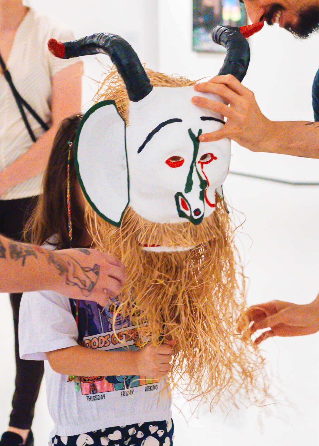 Descrição da imagem: Fotografia vertical de uma menina vista de frente, em pé. Ela é branca, de cabelos lisos castanho-escuros. Usa camiseta branca com estampa dos personagens do filme divertida mente.
Mão masculina segura diante do rosto dela uma grande máscara — uma das obras de Zimar. A máscara tem chifres pretos voltados para trás, com as pontas vermelhas voltadas para cima. Atrás dos chifres, cabelos lisos feitos de palha marrom-clara. O rosto da máscara é branco, com sobrancelhas pretas finas e arqueadas. Os olhos são vazados e contornados por delineado vermelho. O nariz é verde e irregular. As orelhas, grandes e pontiagudas, são brancas, com uma linha verde nas bordas. Os lábios são vermelhos. O bigode e a barba, lisos, também são feitos de palha marrom-clara. Fim da descrição da imagem.
