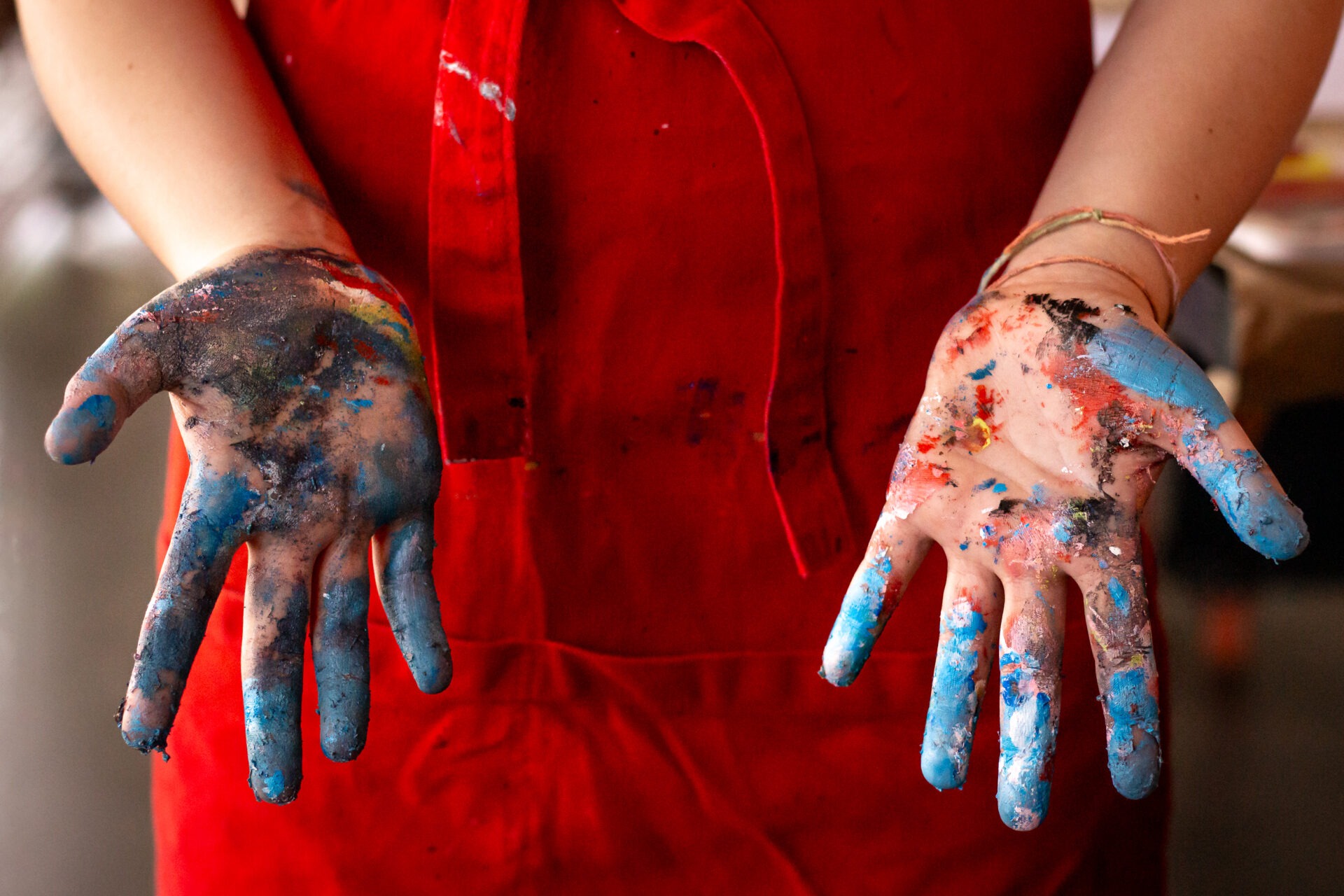 Descrição da imagem: Fotografia horizontal de mãos sujas de tinta. Vistas de perto, até os antebraços, duas mãos estão abertas e posicionadas à frente, com as palmas voltadas para cima. As mãos estão cobertas por manchas de tinta em cores como azul, vermelho, preto e amarelo. No punho esquerdo, há pulseiras finas de pano. A pessoa está com um avental vermelho. Fim da descrição da imagem.