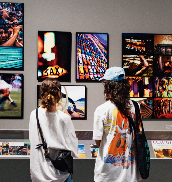 Descrição da imagem: Fotografia vertical de duas pessoas vistas de costas observando fotografias coloridas emolduradas, dispostas em uma parede cinza. A pessoa à esquerda é branca, de cabelos castanhos longos e ondulados, com mechas loiras, presos em um coque baixo. Usa camiseta branca larga, shorts jeans e uma bolsa preta a tiracolo. A pessoa à direita é branca, de cabelos longos e ondulados em corte V. Usa boné azul, camiseta branca larga com desenho colorido nas costas, bermuda jeans e uma ecobag com a alça sobre o ombro direito. As fotos na parede exibem cenas urbanas, entre elas jogo de futebol, pessoas e carros. À frente da parede, um balcão baixo com tampo de vidro exibe cartões e revistas dispostos lado a lado. Fim da descrição da imagem.
